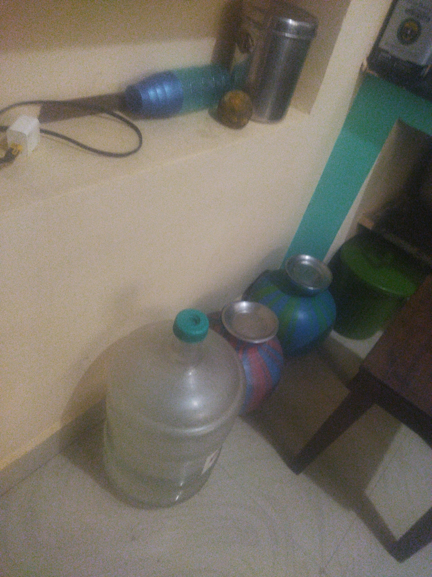 An indoor scene, likely a room, displays a collection of water containers and other items on a floor and shelf. In the foreground, a large, clear plastic water jug with a turquoise cap is partially visible. To its left and slightly behind, two round, blue and striped containers are stacked, each topped with a metal lid. Further back and to the right, a dark green plastic bucket stands. On a shelf above and to the left, a blue water dispenser with a black cord is positioned next to a metal canister. A small, light-colored object, possibly a power adapter, is connected to the cord of the water dispenser. A small, golden-brown spherical object rests near the water dispenser. The walls are a light beige color, and a portion of a teal-colored vertical stripe is visible on the back wall. The flooring appears to be a light gray, tiled surface. Shadows suggest lighting from the right side of the scene.