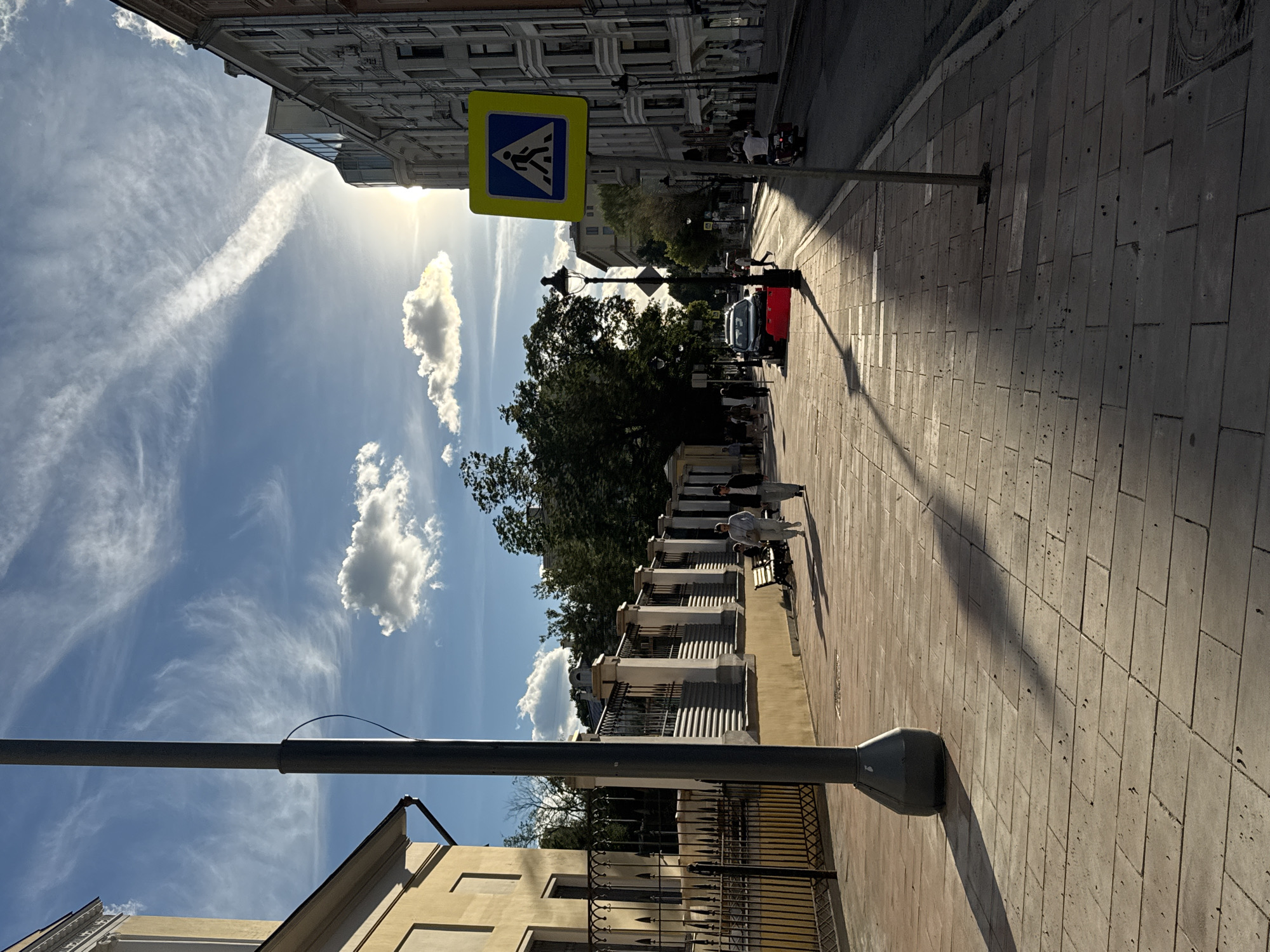 The image depicts a street scene in Moscow, Russia, bathed in the bright sunlight of a clear day. The perspective is slightly tilted, giving a dynamic feel to the composition.
Dominating the left side of the frame is a building with multiple windows and architectural details, suggesting an urban environment. Above, a vibrant blue sky is adorned with wispy white clouds and a prominent contrail, indicative of an airplane passing overhead. The sun is high, casting long shadows across the scene, suggesting it is either late morning or early afternoon.
A pedestrian crossing sign, a yellow square with a blue diamond containing a white silhouette of a person walking, is prominently displayed on a pole. This sign, along with the paved sidewalk and the presence of people, confirms the setting as a public urban area.
Several people are visible on the sidewalk, walking and one person appears to be sitting on a bench. A red car and a scooter are also present on the street, indicating ongoing activity and traffic. The architecture of the buildings on the right side of the street mirrors the style of the building on the left, reinforcing the impression of a historic urban setting. The ground is paved with rectangular stone tiles, adding to the textured appearance of the scene.
A street lamp stands tall in the foreground, its dark silhouette contrasting with the bright sunlight. The overall atmosphere is one of a pleasant, active day in a city. No visible text is discernible in the image.
