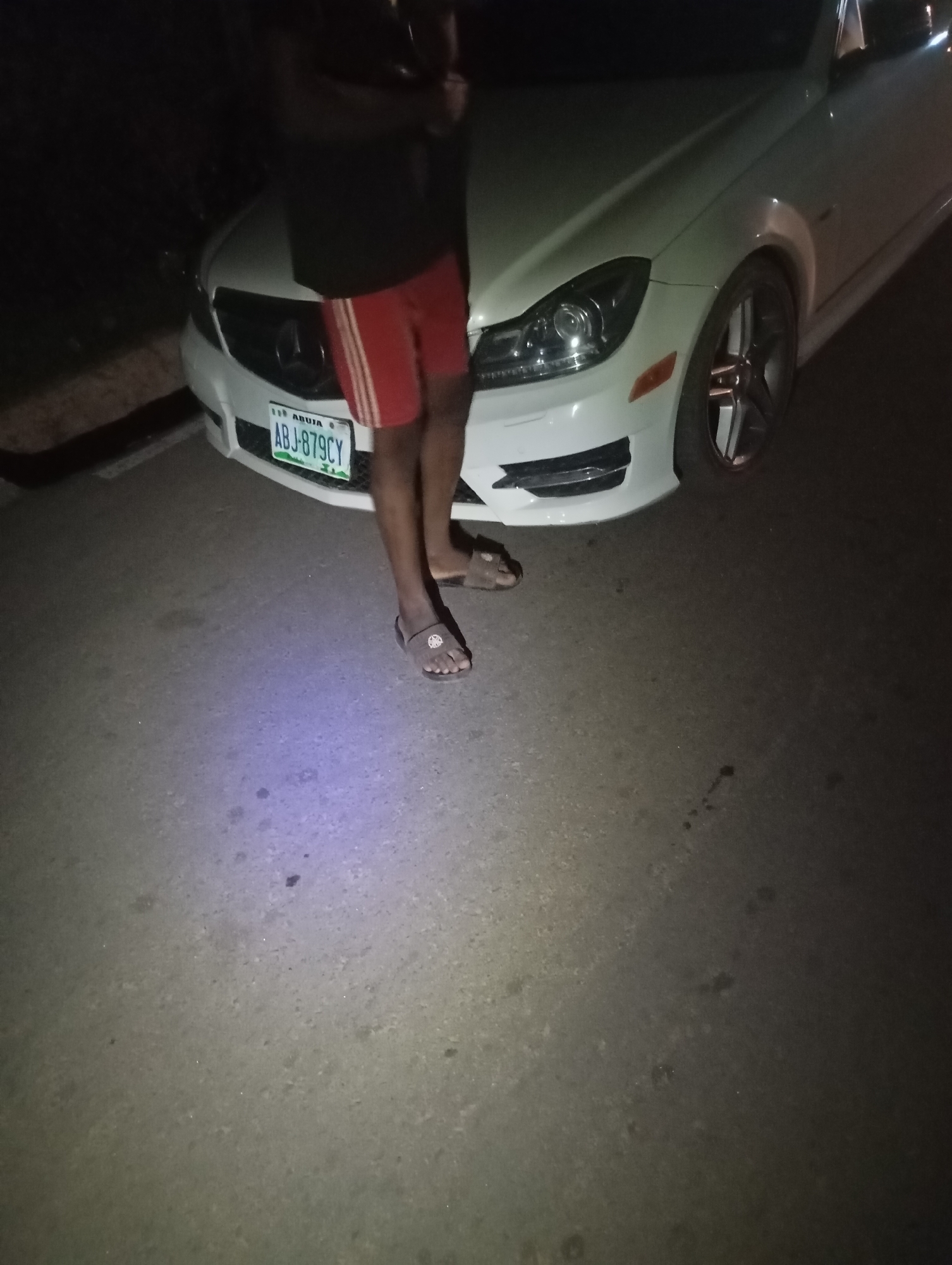 A person is standing next to a white Mercedes-Benz C-Class, license plate ABJ-879CY, with red and white striped shorts and dark sandals. The scene is outdoors at night, with the ground illuminated by a purple light source. The car is parked on a paved surface, possibly a road or driveway. The person appears to be leaning against the car.