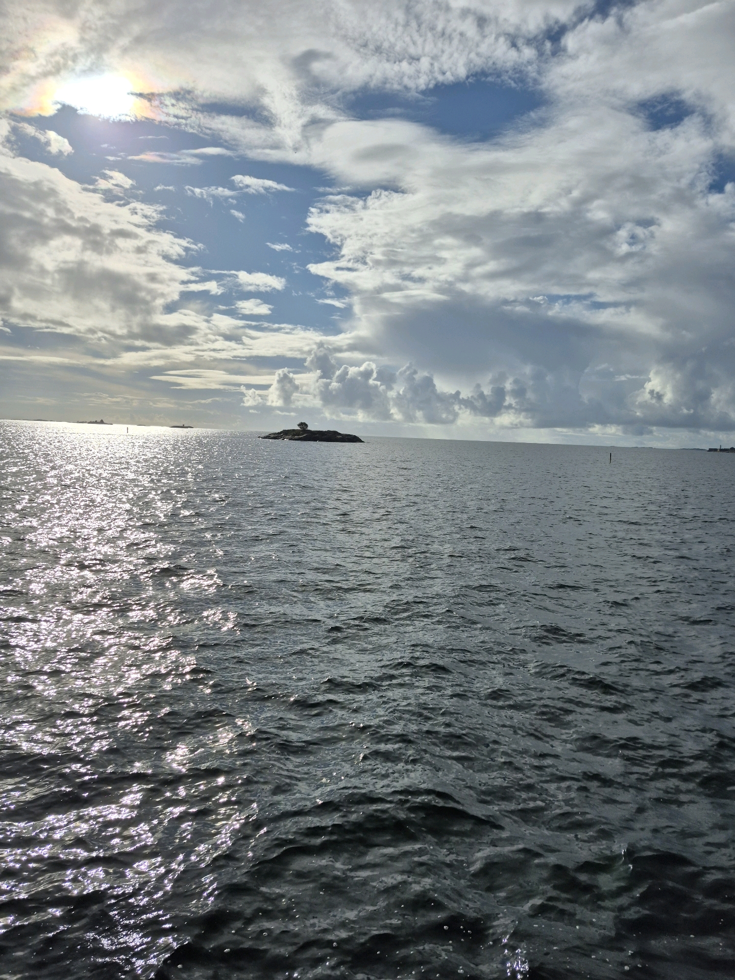 The image depicts a serene marine environment with a cloudy sky overhead and the sun partially visible, casting a shimmering reflection on the water's surface. In the distance, a small, tree-covered island is visible, adding a touch of natural beauty to the scene. The sea is calm with gentle ripples, and several distant vessels and buoys can be seen on the horizon, suggesting maritime activity. The overall atmosphere is tranquil and expansive, evoking a sense of peace and solitude.
This image was taken in Hanko, Finland. The sun's halo effect, visible in the upper left corner, is a meteorological phenomenon that can occur when sunlight interacts with ice crystals in the atmosphere.