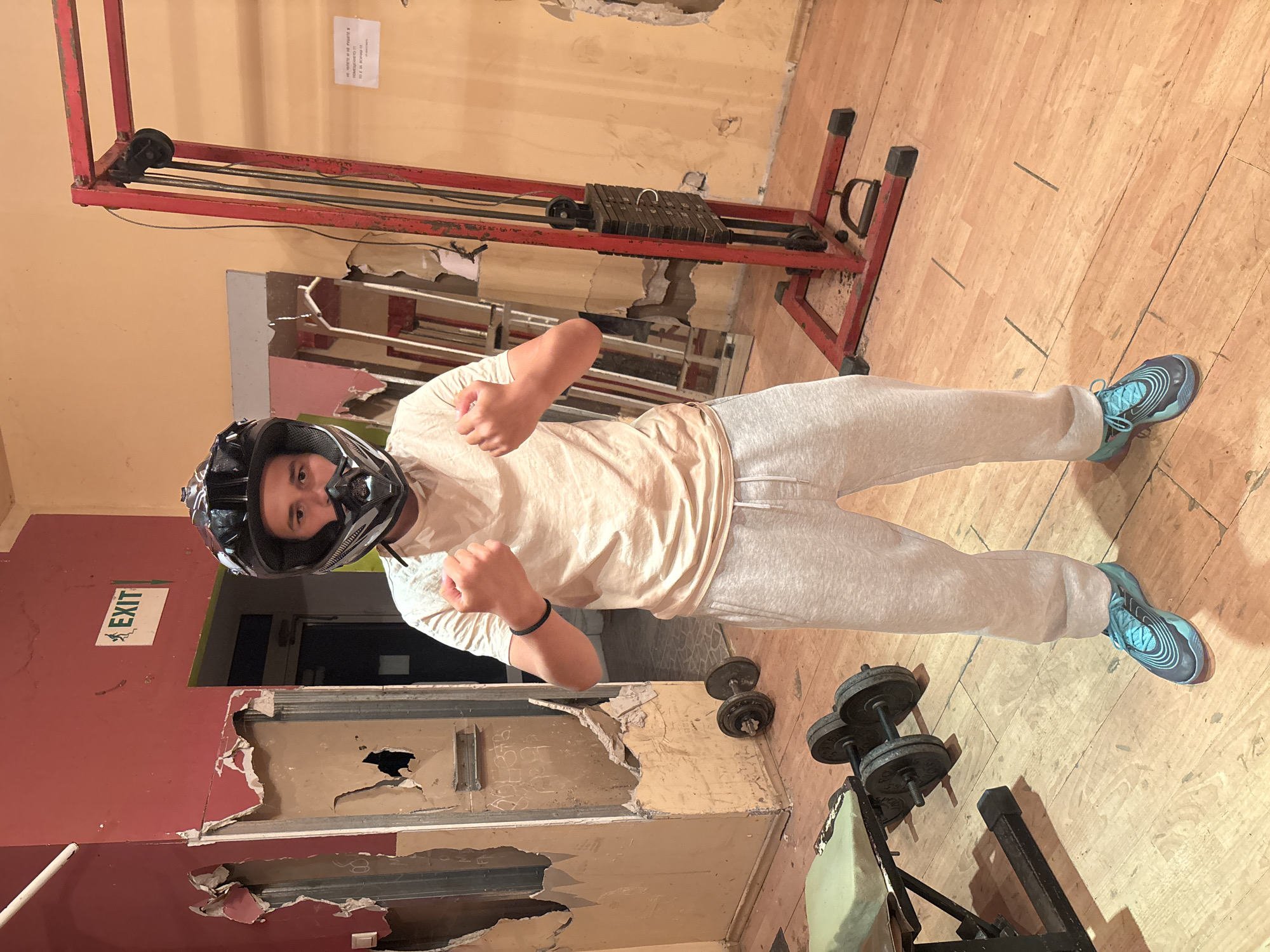 A person wearing a motorcycle helmet, a t-shirt, and sweatpants stands in a room that appears to be a poorly maintained gym. The person's fists are raised in a boxing stance. Behind the person is a doorway with an "EXIT" sign above it. The walls are peeling and damaged. Gym equipment, including a weight machine and dumbbells, is visible. The location is Goritsa, Bulgaria.