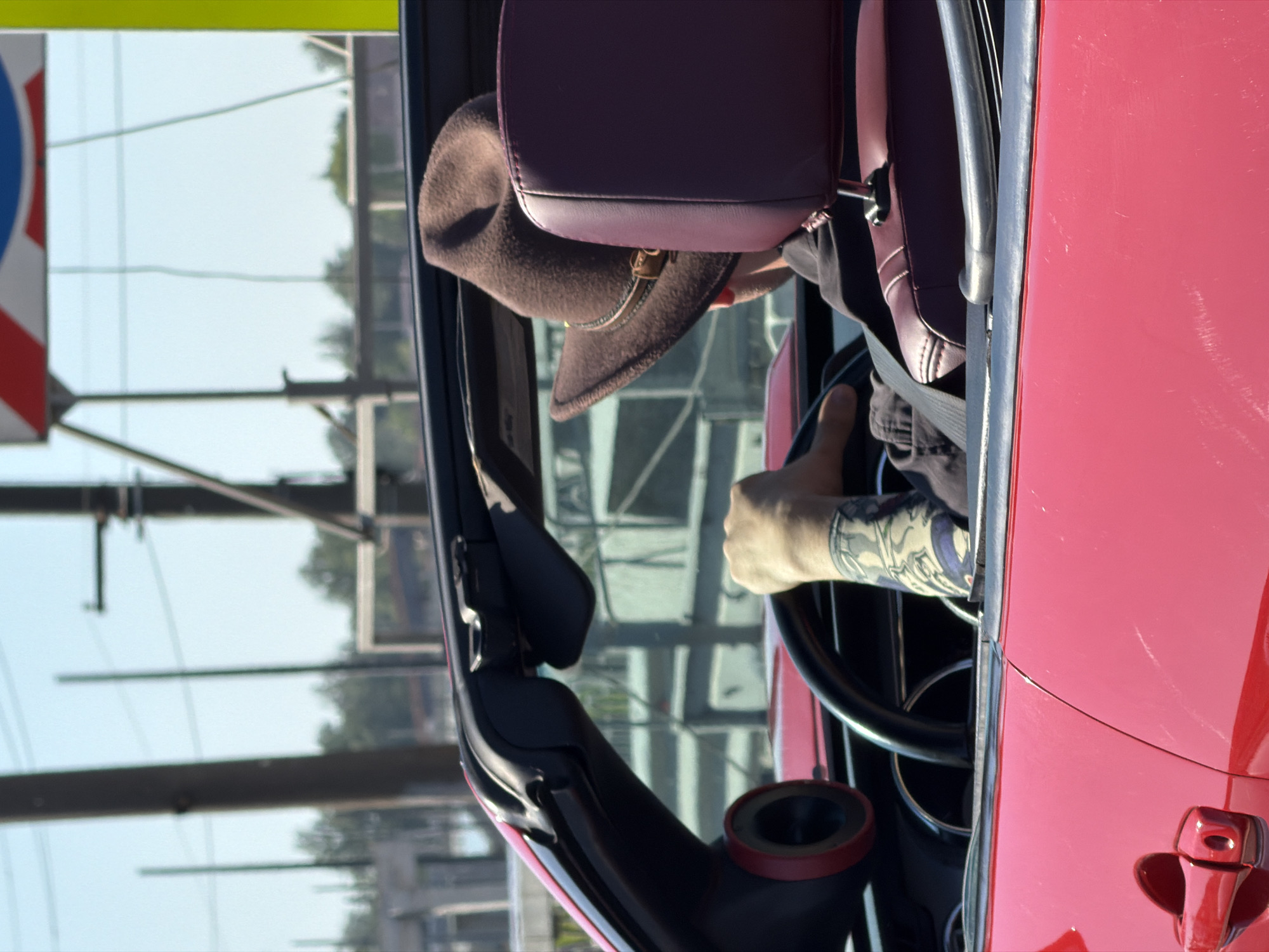 The image shows the interior of a red convertible car. A person's hand, tattooed, is gripping the steering wheel. A brown hat sits on the back of the passenger seat. Outside the car window, power lines and a structure with vertical stripes are visible, suggesting an urban environment. Considering the presence of the structure with vertical stripes, it is possible that the location is Saint Petersburg, Russia, as these structures are often associated with road signs or urban markers in that city.