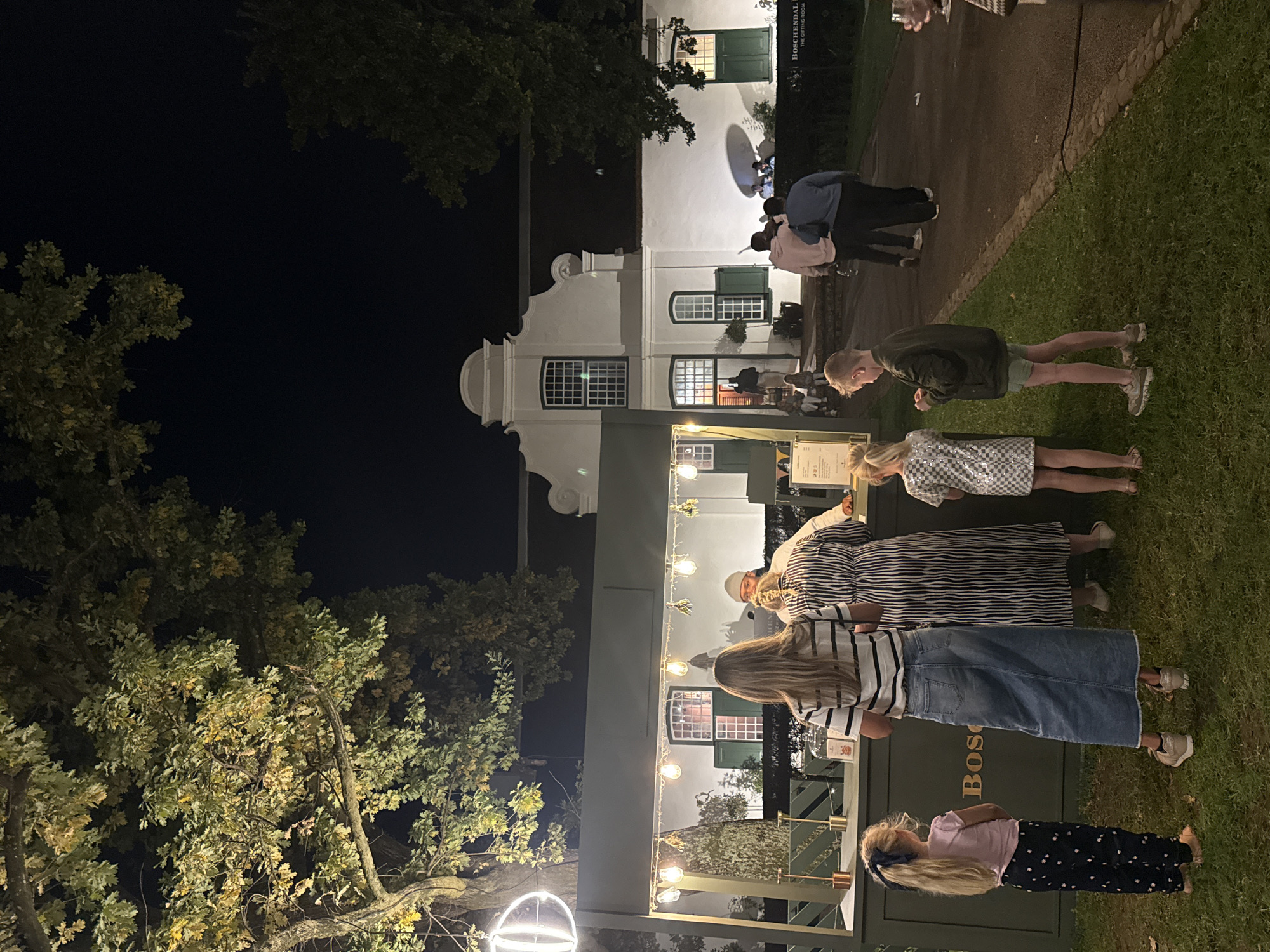 An outdoor, nighttime scene depicts a social gathering at the Boschendal estate in Stellenbosch, South Africa. The background is dominated by a majestic white Cape Dutch style building, featuring classic gables and prominent green window shutters. A sign on the building clearly reads "BOSCHENDAL" and "THE GIFTING ROOM".
In the mid-ground, a green-painted stall, also bearing the "BOSCHENDAL" name, is illuminated by decorative string lights with exposed bulbs. Several people are congregated around this stall, suggesting it might be a food vendor or market stand. To the left, large trees with autumnal or illuminated foliage frame the scene against a dark night sky.
A diverse group of individuals, including adults and children, populates the foreground and mid-ground. In the immediate foreground, a young girl with blonde hair, wearing a pink top and dark patterned pants, stands with her arms crossed. Adjacent to her, a woman with long blonde hair, dressed in a striped t-shirt and a long denim skirt, is seen looking towards the stall. Further back, another person in a striped dress and a young boy in a dark jacket and shorts are visible. Other figures are scattered throughout the scene, some appearing to be conversing or queuing near the building.
The scene is lit by artificial lights, creating a warm and inviting ambiance against the dark of night. The presence of grass and a paved path indicates an outdoor setting. The overall impression is one of a relaxed evening event or market at a historic wine estate.