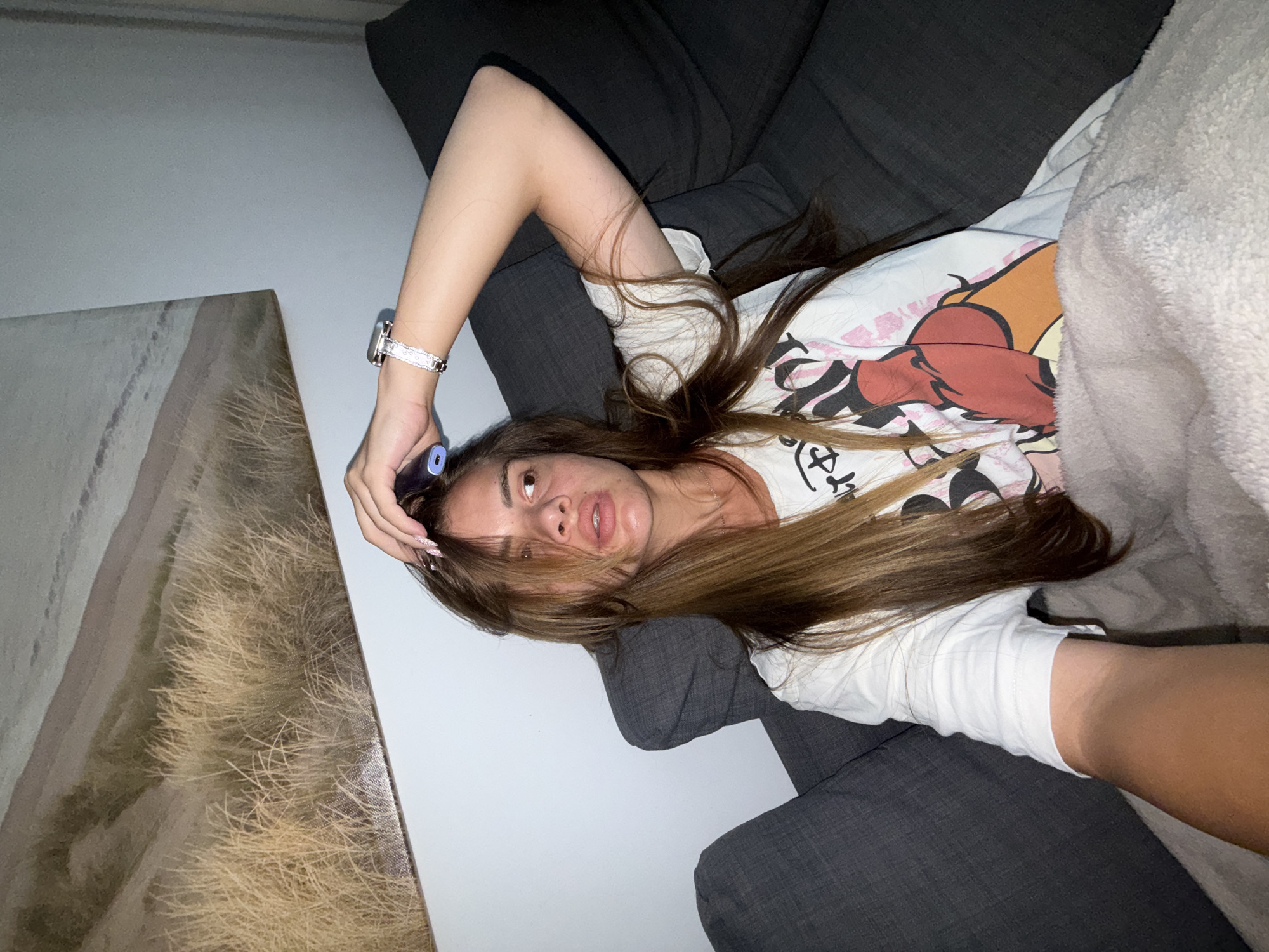 A woman with long, brown hair is lying on a dark gray sofa, partially covered by a beige blanket. She is wearing a white t-shirt with a graphic print. Her right hand is raised to her head, and she is wearing a silver watch with a patterned band on her left wrist. She is looking to the right, away from the camera. Behind her, on the wall, is a large framed artwork depicting a landscape with dry grass and sand dunes. The room is dimly lit, creating shadows on the sofa. There is no indication of the specific location within the city of Plovdiv, Bulgaria, as the image depicts an indoor scene with no identifying landmarks.