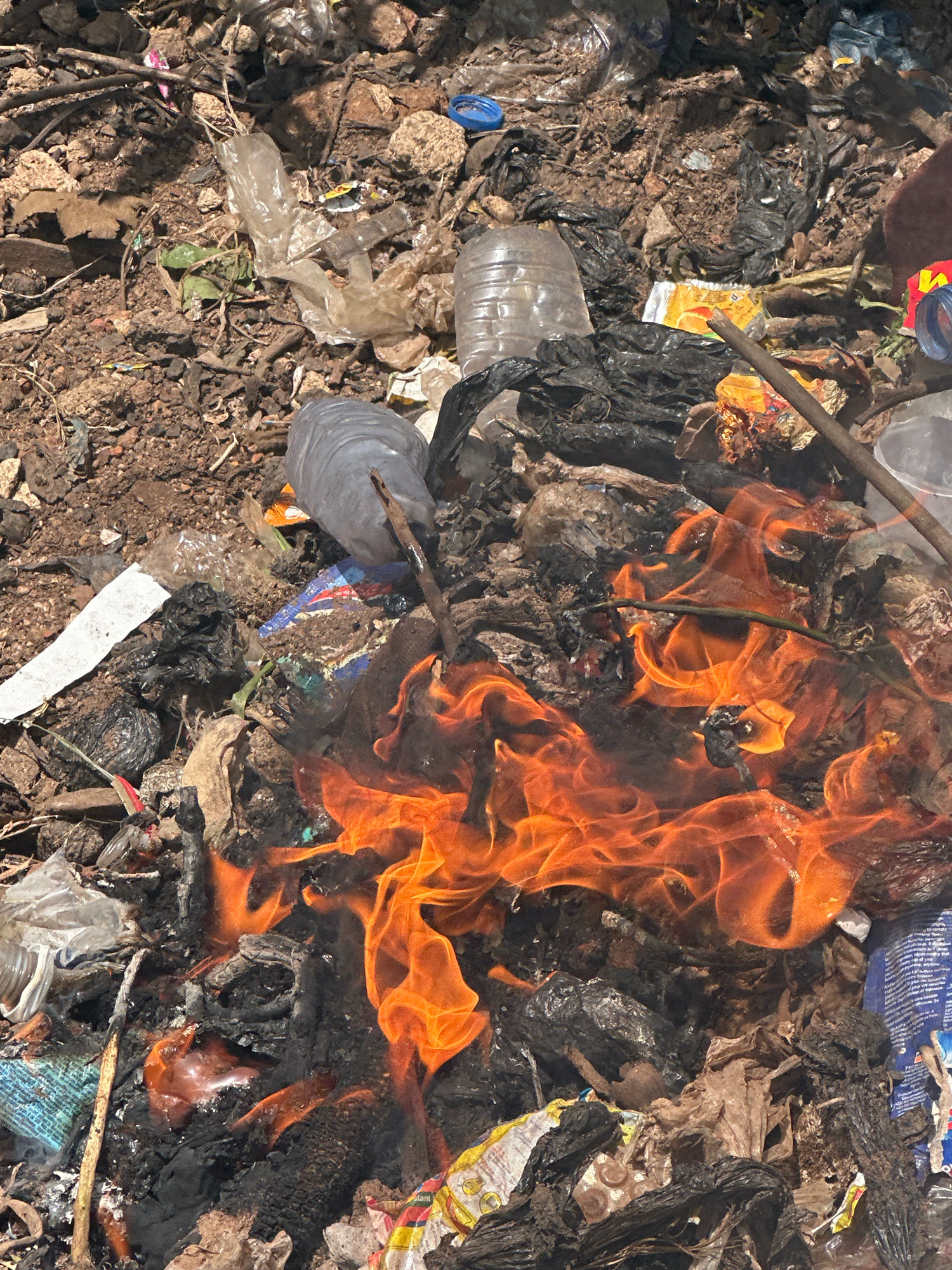 This is a close-up, overhead shot of a small fire burning in a pile of garbage. The fire is concentrated in the center of the frame, with vibrant orange flames consuming various types of waste. The surrounding debris consists of plastic bottles, black plastic bags, torn packaging, and dry organic matter, all mixed with dirt and soil. Some of the plastic appears to be partially melted and discolored from the heat. The overall impression is one of an uncontrolled burning of refuse. The scene is located in Gura Top, Nigeria.