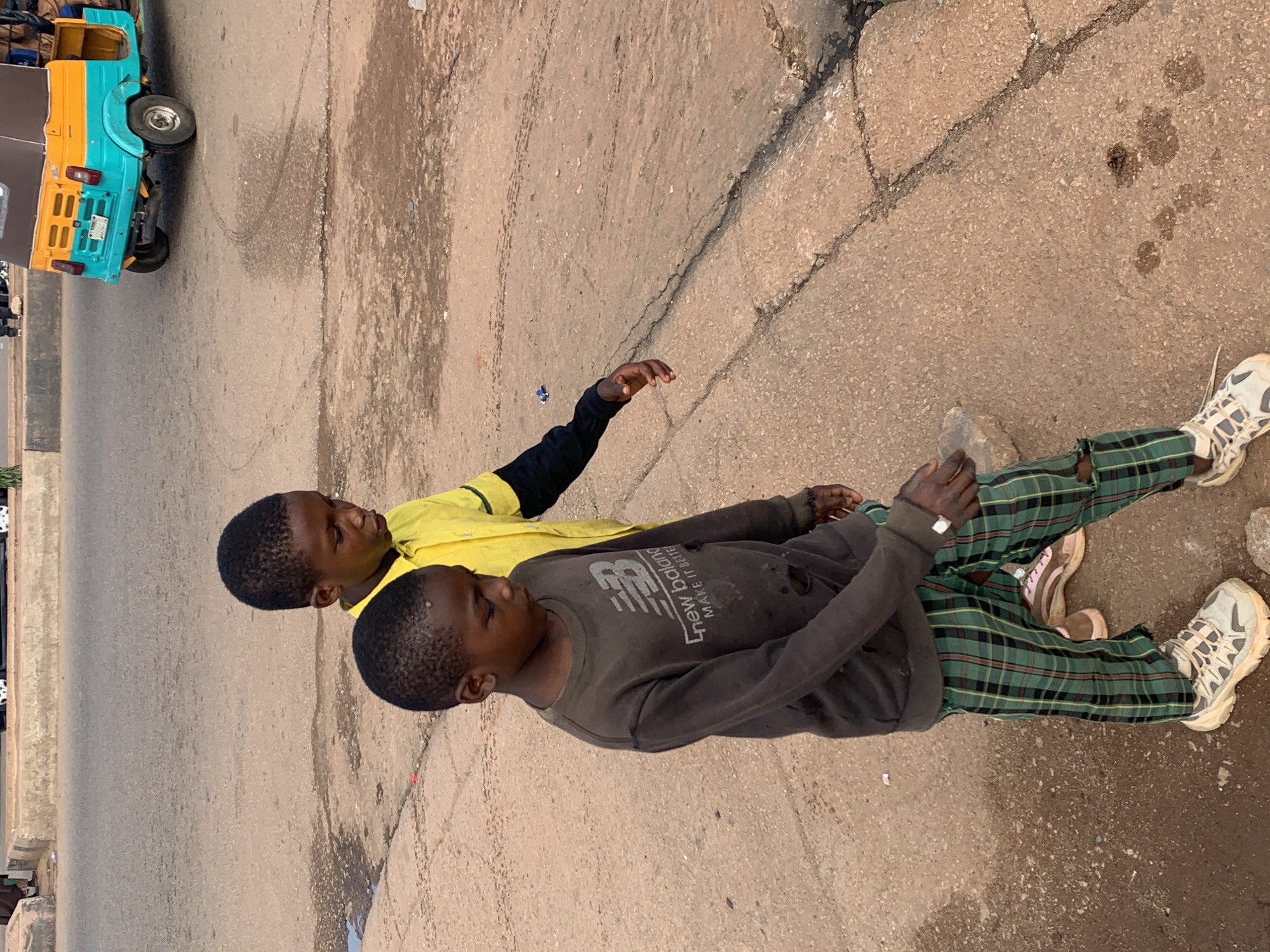 Two young boys are walking on a paved road in Gura Top, Nigeria. The boy in the foreground is wearing a dark grey long-sleeved shirt with a "New Balance" logo and ripped plaid pants in green and black. He is also wearing light-colored athletic shoes. The boy behind him is wearing a bright yellow long-sleeved shirt with black sleeves. They are walking side-by-side. In the background, a yellow and turquoise auto rickshaw is parked on the road. The road surface is cracked and worn, with some areas of wetness and debris. The environment appears to be an urban or semi-urban setting.