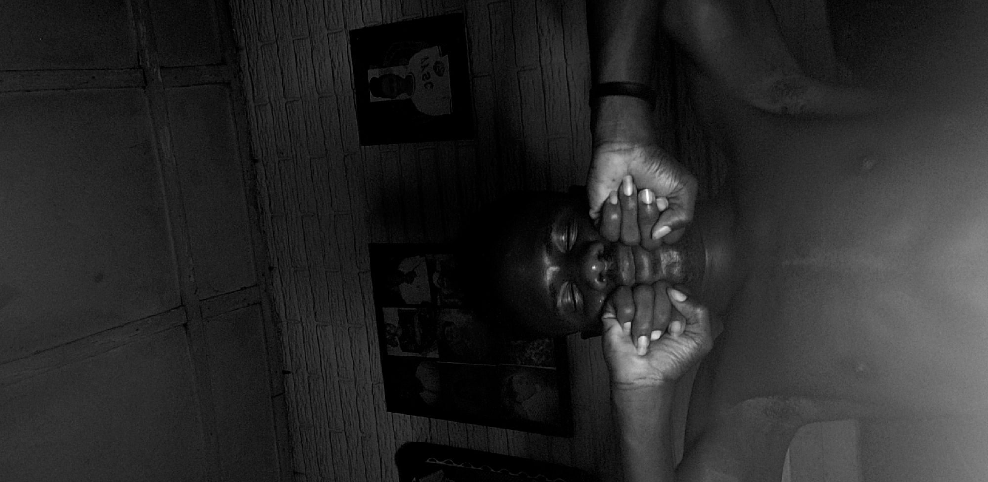 A black and white photo shows a person lying down with their hands pressed against their face, almost as if obscuring it. The person is dark-skinned. They have a bracelet on one of their arms. The background includes a brick-like wall with framed pictures. A part of the room's wooden structure is also visible. The overall scene is low-lit and appears to be indoors. The location is Narayi, Nigeria.