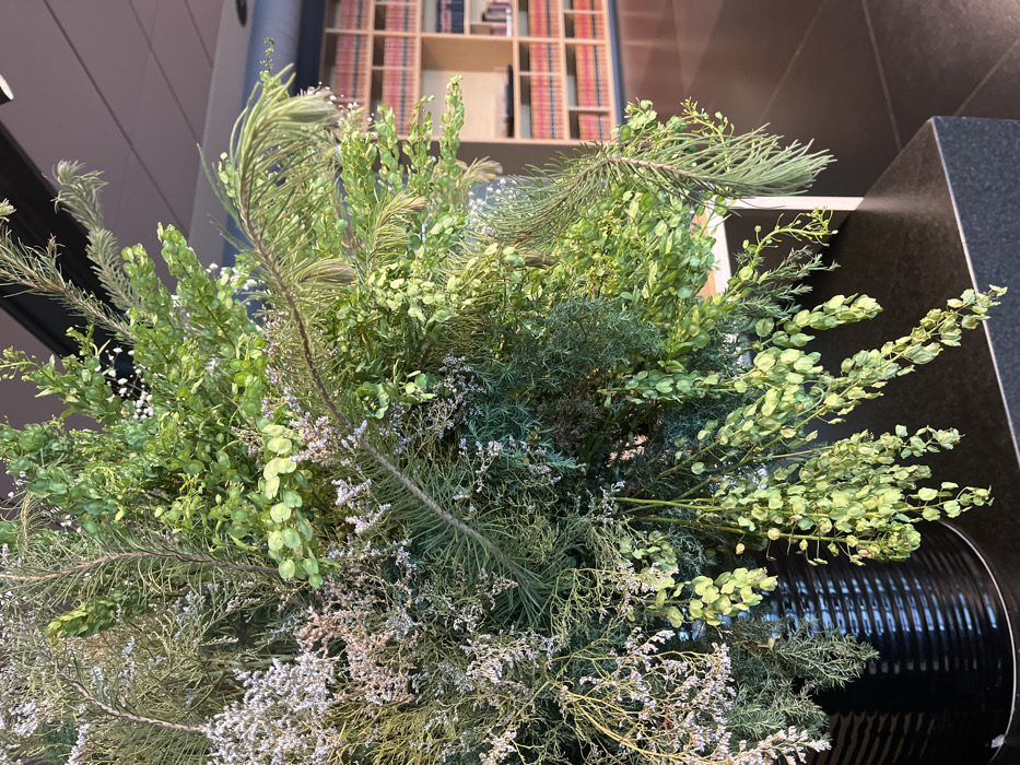 An indoor scene in Mexico City features a prominent, large, mixed-foliage botanical arrangement positioned in the foreground. The arrangement, contained within a black, vertically ribbed cylindrical planter, consists of diverse green plant material including needle-like branches, broad ovate leaves, and lighter, dried, wispy flora. This display rests on a dark, flat, possibly stone countertop or ledge. In the background, a multi-tiered system of light-colored wooden bookshelves is visible, filled with numerous books predominantly featuring red-orange spines, indicating a library or substantial book collection. The floor appears dark, and the ceiling is dark with integrated lighting. No people are present. The precise location within Mexico City is not discernible from the visual evidence.