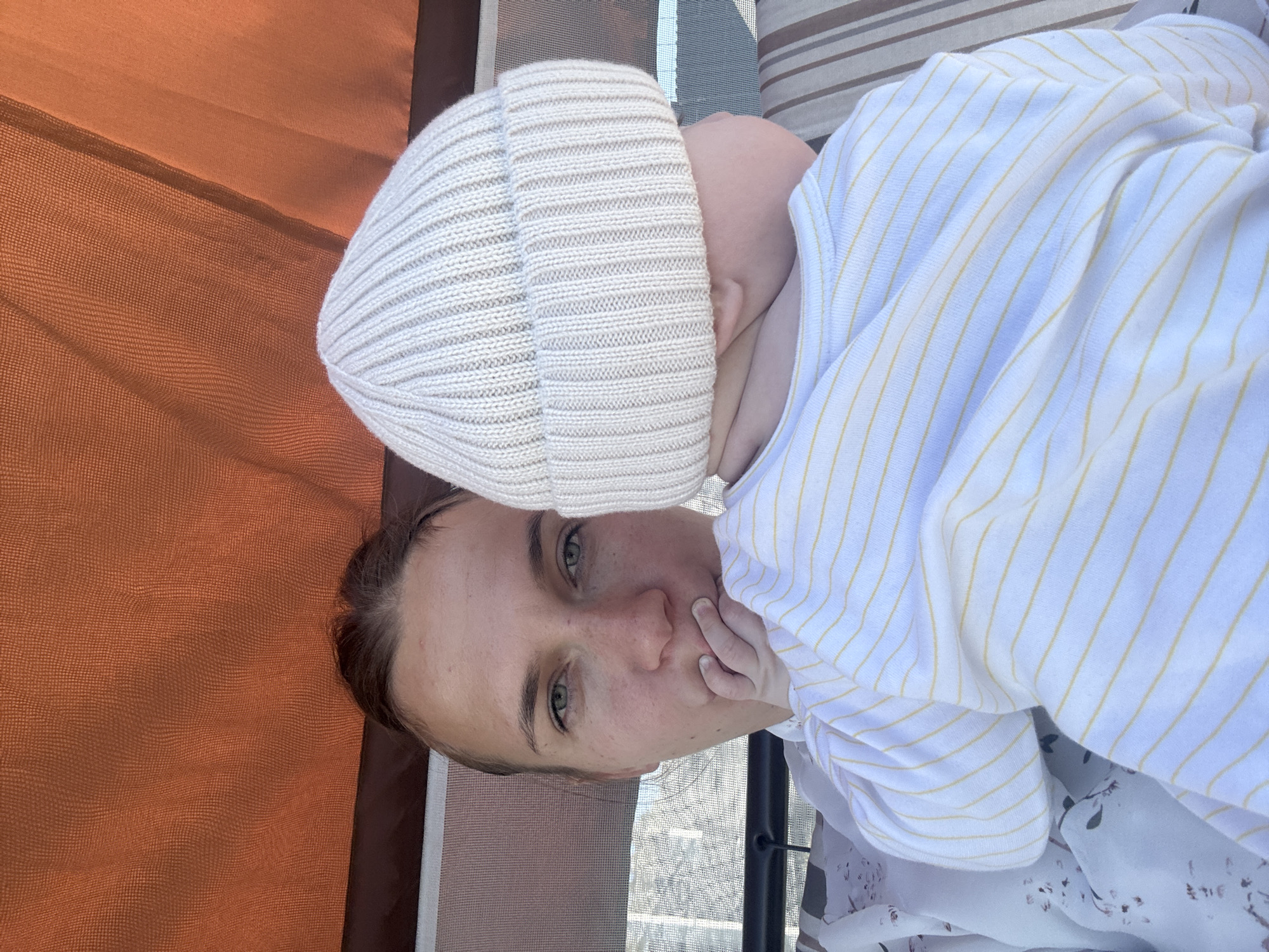A woman is lying down, holding a baby who is wearing a white ribbed beanie and a white shirt with yellow stripes. The woman is fair-skinned with green eyes and her hair is pulled back. The background includes a striped surface, a window-like grid, and an orange curtain. The location is inferred to be Razdolye, Russia, based on the prompt's specifications, but there are no discernible landmarks in the image to confirm this.
