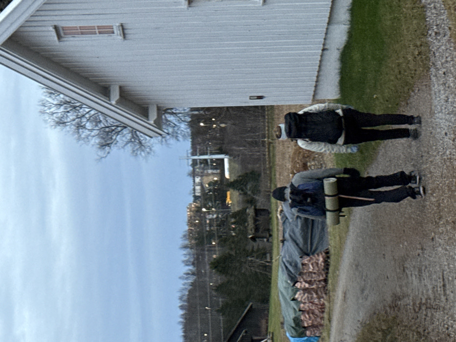 The image depicts two individuals walking away from the viewer on a gravel path in Sand, Norway, under an overcast, light grey-blue sky. To the immediate right, a white, vertical-paneled wooden building with a pitched roof and a visible window frame stands. On the left side of the path, a large, dark-colored tarp covers a row of stacked items, partially secured by multiple camouflage-patterned bags. Further left, a small, dark wooden structure with a moss-covered roof is discernible amidst bare trees. In the distant background, illuminated structures, streetlights, power lines, and a distinct white pointed spire indicate a town.
The lead individual, positioned to the left on the path, wears a dark beanie, a grey long-sleeved top, dark trousers, dark gloves, and a blue backpack with a tan rolled mat horizontally strapped beneath it. The second individual, on the right, wears a light-colored jacket, dark trousers, a light-colored headband or ear warmer, possibly light blue gloves, and a large black backpack. Both individuals are equipped with backpacks, consistent with outdoor travel or hiking.