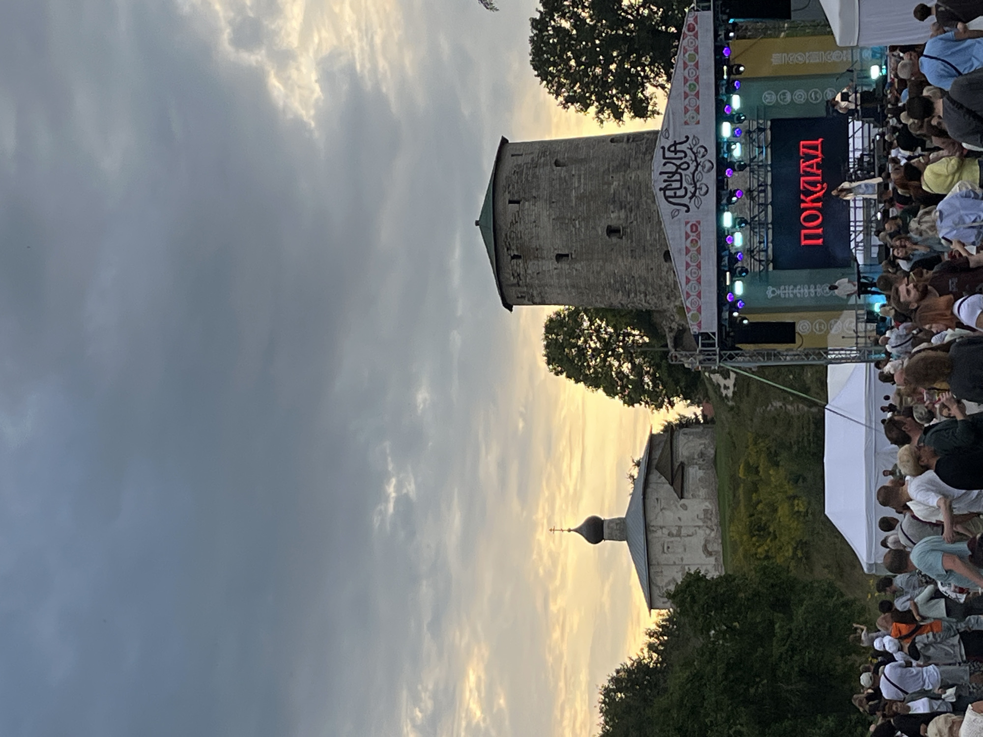 The image shows an outdoor festival or concert at an event space likely located near historical landmarks in Pskov, Russia. In the background, on a hill, stands a medieval stone tower with a green roof and a nearby white, domed church with a cross on top. In the foreground, a crowd of people is gathered in front of a stage. The stage has a large screen displaying the word "ПОКЛАД" in red Cyrillic letters, and lights are illuminating the performers. A white tent is also visible in the crowd. The sky is cloudy with a hint of sunset colors peeking through. The overall scene depicts a cultural event taking place in a historically significant setting.