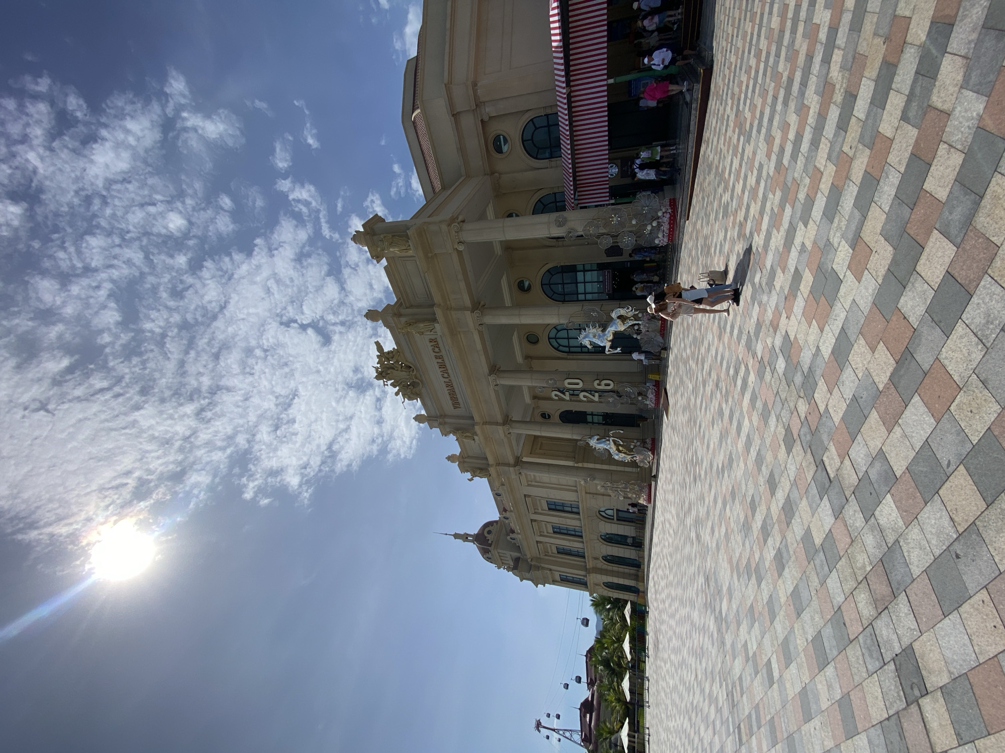 The image shows the entrance to Vinpearl Cable Car in Nha Trang, Vietnam. The building has a grand, neoclassical architectural style with large columns and statues. In front of the building is a large plaza paved with colorful square tiles. Several people are visible in the plaza, some standing and talking, others walking towards the entrance. A large number of white umbrellas are set up in the plaza, indicating a waiting area or outdoor seating. In the background, several cable cars are seen suspended from wires, connecting to a distant structure. The sky is bright blue with scattered white clouds, and the sun is shining, casting shadows on the ground. The scene appears to be a popular tourist attraction on a sunny day.
