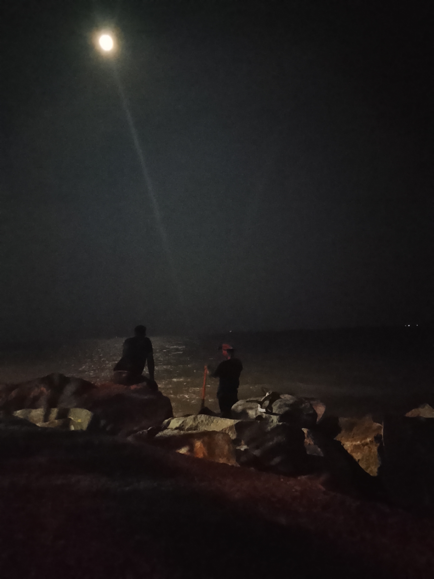 The image depicts a nighttime scene by the sea in Tanjung Balai Karimun, Indonesia, under a full moon. Two individuals are visible. One person is seated on large rocks near the water's edge, facing away from the camera and towards the sea. The second person stands further to the right, also facing the sea and holding a long, thin object, possibly a fishing rod or a pole. The sea reflects the moonlight, creating a shimmering effect. The foreground is dominated by large, dark rocks, suggesting a breakwater or a rocky shoreline. The overall atmosphere is serene and dark, characteristic of a quiet night.