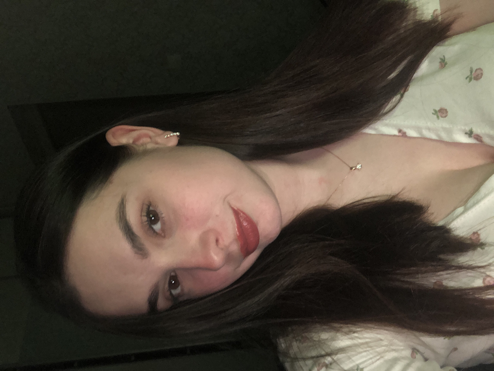 A young woman is lying down with her head tilted towards the camera. She has long, dark brown hair and is wearing a light-colored top with a floral pattern. She is wearing lipstick and a necklace with a small pendant. Her ear is visible and appears to have a piercing. The background is dark and indistinct.
There is no indication of the city of Noviy, Russia.