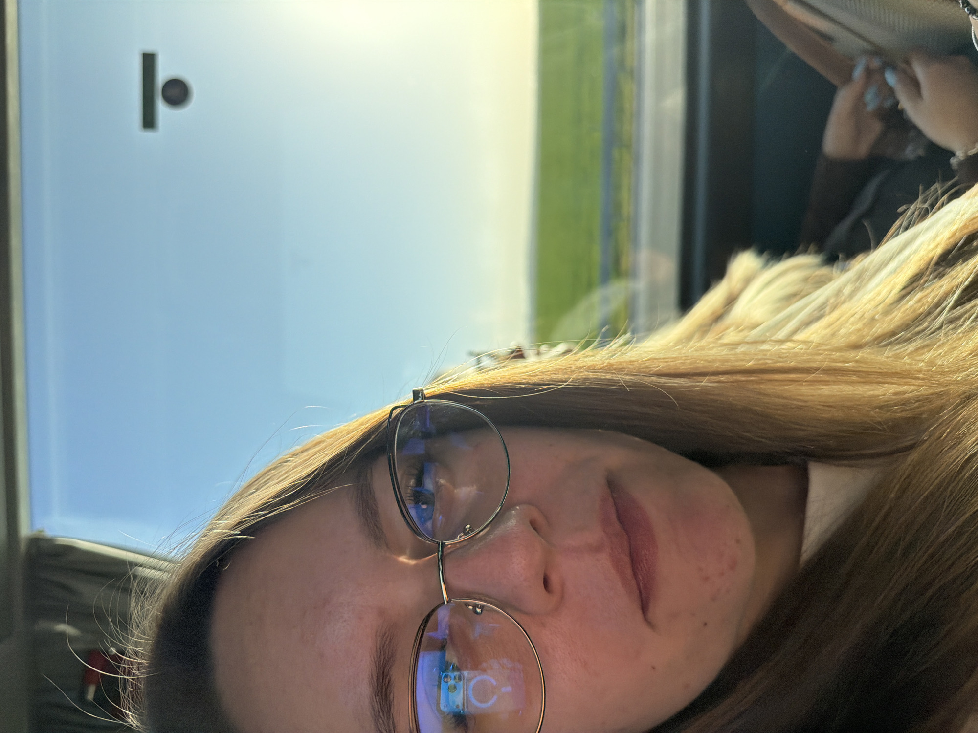 A woman with long blonde hair and glasses is pictured indoors, likely in a vehicle, in what appears to be a selfie. The background visible through the window suggests an outdoor scene with a blue sky and green field. A latch or hook is visible on the upper left of the window frame. The reflection in her glasses hints at interior details, possibly a device screen. Given the specified location of Градина, Bulgaria, it is reasonable to assume the photo was taken within this city or its surrounding area. Another person's hand can be seen in the background.