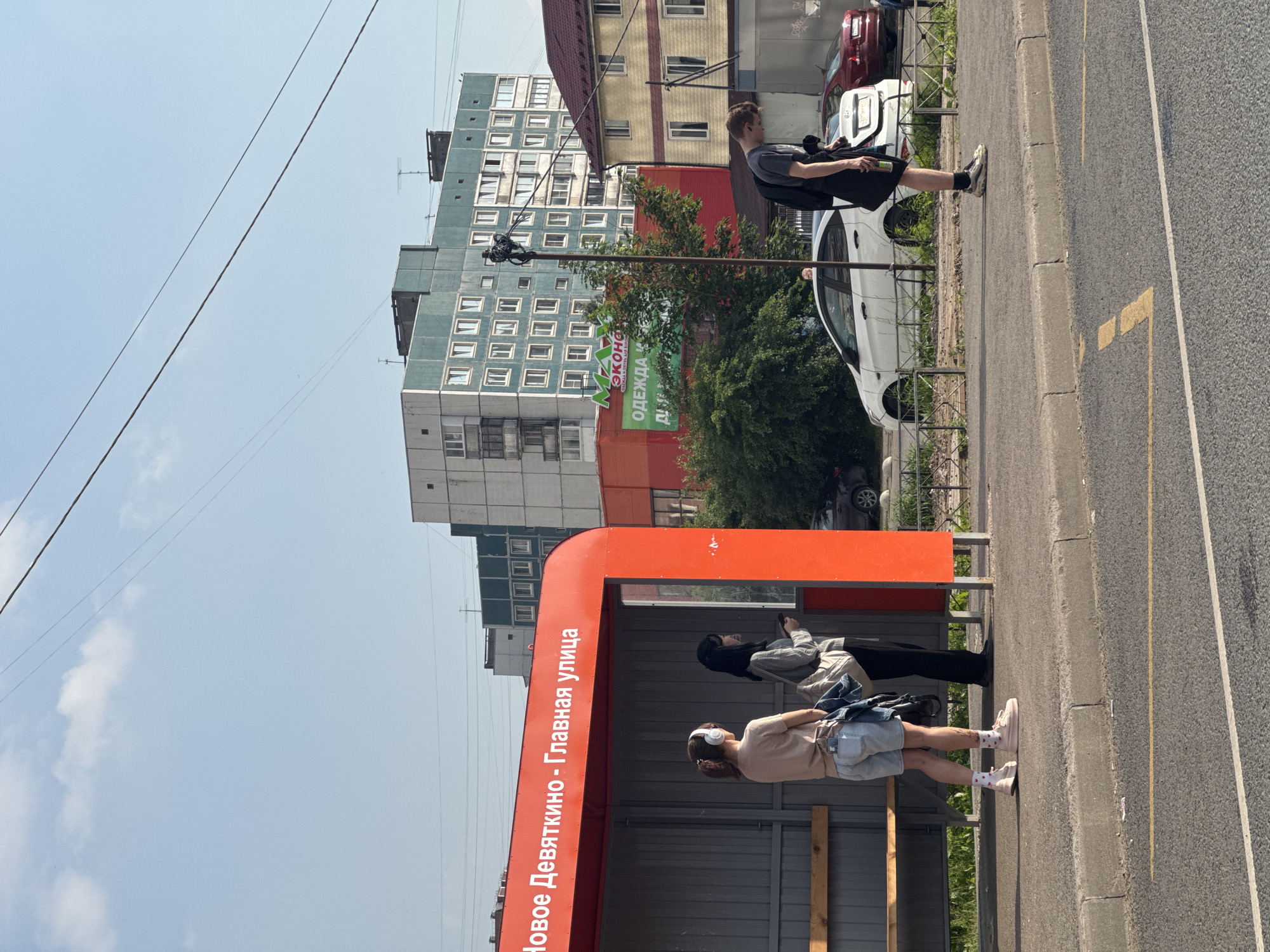 At a bus stop in Novoe Devyatkino, Russia, three individuals are visible. A young woman wearing headphones and light-colored shorts stands on the left, facing away from the camera. Next to her, a woman in a gray sweater and black pants holds a phone and a bag, also facing away. Further to the right, a young man in a gray t-shirt and black shorts stands with his head bowed, holding a bottle. Behind them, buildings and a street with parked cars are present under a clear sky. The bus stop shelter is bright orange.