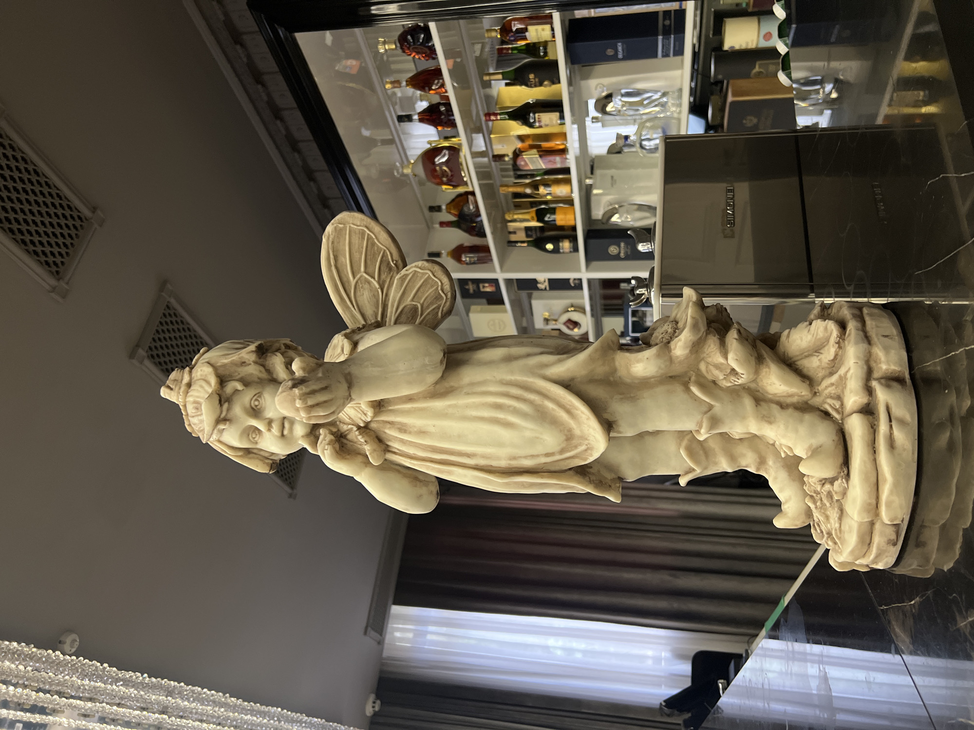 A cream-colored statue of a winged cherub or angel, possibly made of marble or alabaster, is the central subject of this indoor photograph. The statue is depicted in a reclining position, with its head slightly raised and hands clasped near its chin, gazing forward with a serene expression. Intricate wings adorn its back.
The statue rests on a highly reflective, dark surface, likely a polished marble or glass countertop, which mirrors parts of the room. Adjacent to the statue, on this surface, is a black rectangular box with the word "SIMONE" faintly visible on its side.
In the background, a large, well-stocked display cabinet is visible, filled with numerous bottles of what appear to be high-end alcoholic beverages, including cognacs, whiskies, and wines, arranged on multiple shelves. Some bottles are in ornate decanters, and others are accompanied by branded packaging or glassware.
To the left of the frame, two rectangular, decorative grates are installed on a light-colored wall, possibly air vents. Further left and below, a window covered by curtains is partially visible, suggesting an external view is present but obscured. Above the statue, a magnificent chandelier, composed of countless small, glittering crystal or glass elements, hangs from the ceiling.
The overall setting suggests an opulent and sophisticated indoor environment, possibly a bar area, a private lounge, or a lavish living room, characterized by elegant decor and a collection of fine spirits. The lighting appears to be artificial, casting a soft glow. No specific activity is taking place; the image captures a static display. The location is indicated as Yerevan, Armenia.