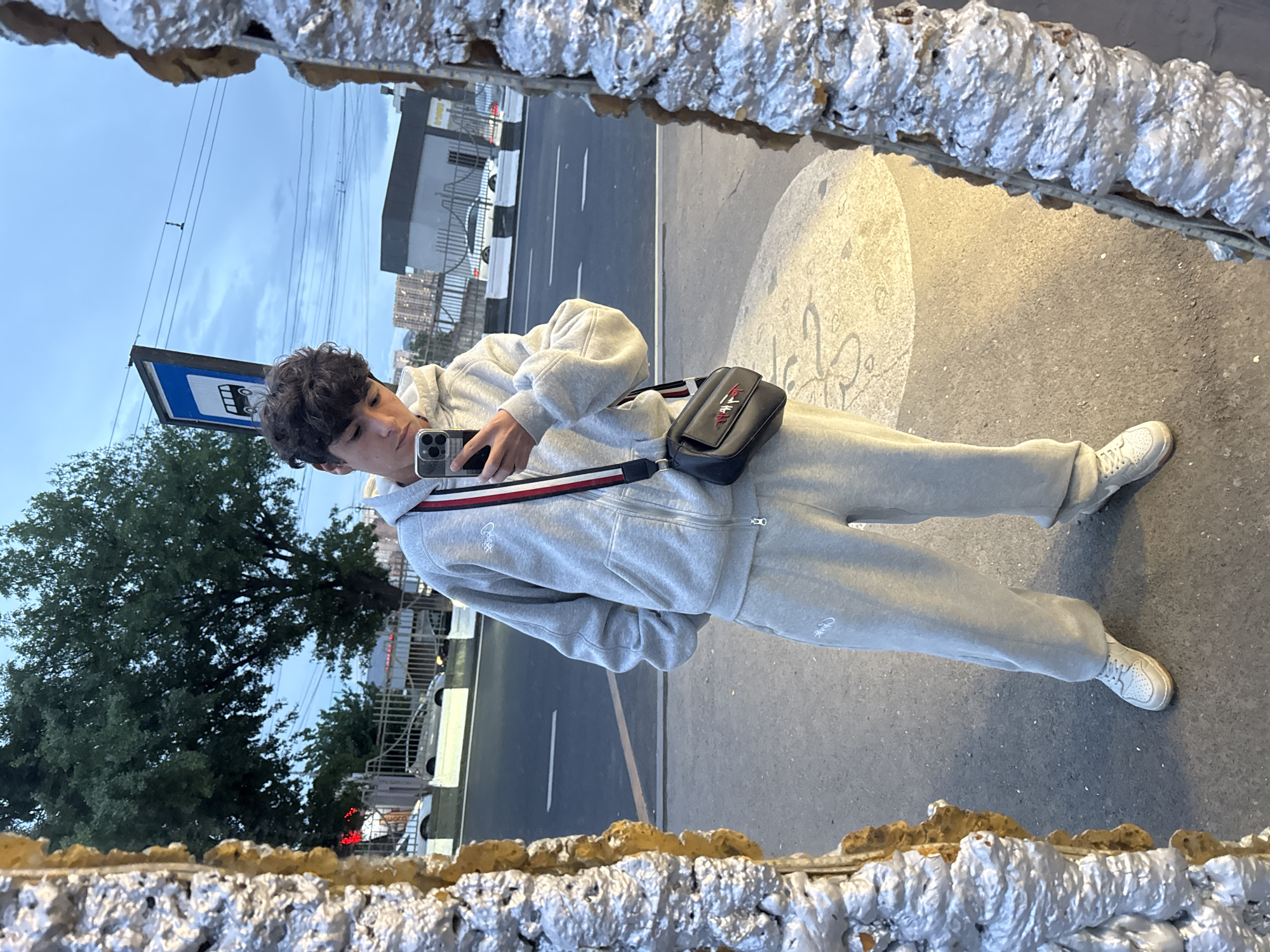 A young man is taking a selfie in a mirror in Yerevan, Armenia. He is wearing a light gray sweatsuit, white sneakers, and a black shoulder bag with a red and white striped strap. The mirror is framed with an ornate, metallic-looking border. The reflection shows a street scene with a bus stop, trees, buildings, and cars. The sky is overcast. The ground is paved.