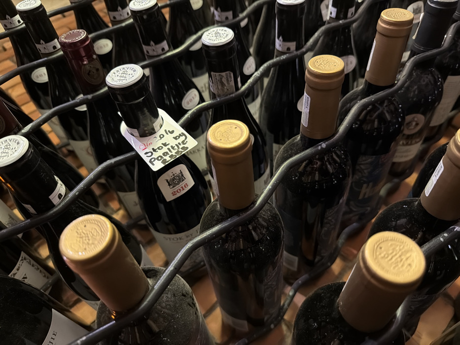 The image displays a close-up, angled view of numerous wine bottles meticulously arranged horizontally within a dark, metallic wire wine rack. The scene is located in a cellar or dedicated wine storage area within Drakenstein Nu, South Africa. The bottles are predominantly dark-colored glass, many visibly coated with a fine layer of dust or sediment, indicating prolonged storage. Varying foil capsules seal the bottle necks: some are bright white or silver, while others are a dull tan or golden hue, several featuring an embossed crest or design. One central bottle, identifiable by a white main label depicting a crest and the year "2016," has a handwritten white tag attached to its neck. The tag displays red handwritten text: "Jan 2016 Putok by Paaltje R&&&". The bottles are cradled individually by the wavy, U-shaped sections of the rack. In the background, a warm, orange-brown brick wall is partially visible through the gaps in the rack and bottles, providing a rustic backdrop to the collection. No individuals are present in the scene.