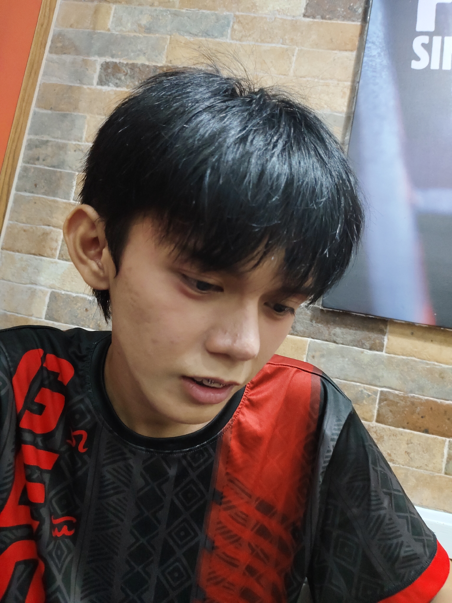 A young Filipino male with dark hair and braces is looking down and to the right. He is wearing a black t-shirt with red accents and a geometric pattern. He has a bruise under his left eye. The background consists of a textured brick wall and a portion of a poster with white text on a dark background. The scene is likely indoors, possibly in a commercial establishment, given the poster and the lighting. This image does not contain enough information to definitively place it in Davao City, Philippines, or any specific city.