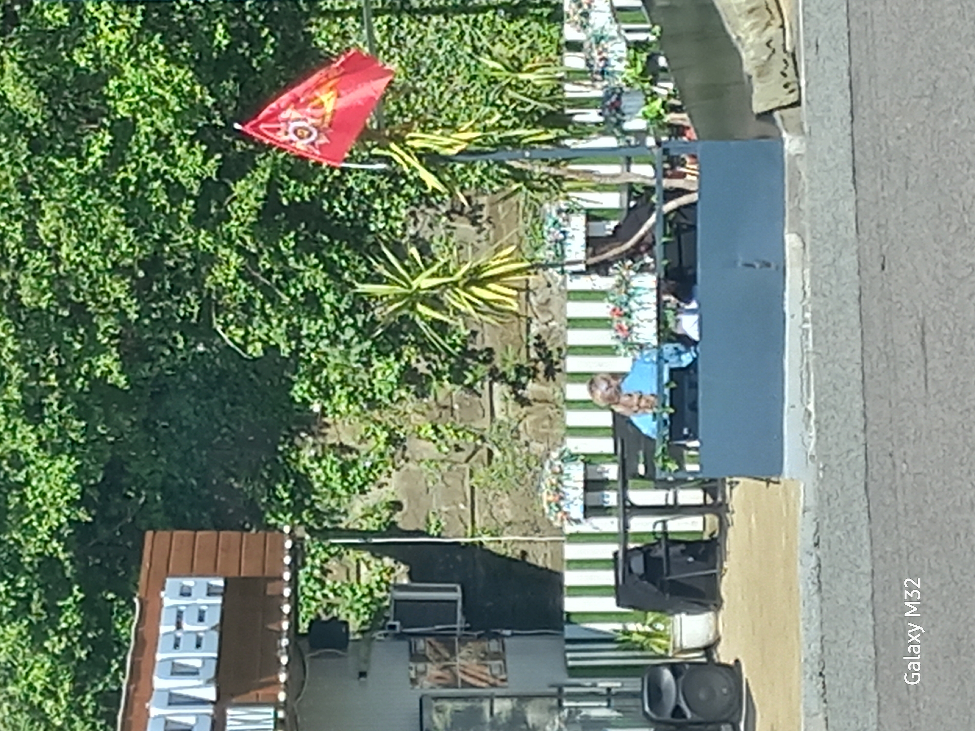 The image depicts an outdoor scene in Sochi, Russia. A red flag, decorated with a golden emblem, is planted amidst dense green foliage. Next to the flag, there are several plants resembling agave. Adjacent to the vegetation is a white picket fence with occasional floral decorations. Behind the fence, a person in a blue shirt is partially visible, obscured by a large blue gate that might be made of metal. In the foreground, there's a building with a brown roof and a sign with the word "BEER" in large, silver letters. Near the building, one can see an air conditioning unit, a speaker, and other equipment, possibly related to a business. The scene is captured from an elevated perspective. The edge of a road is also visible.