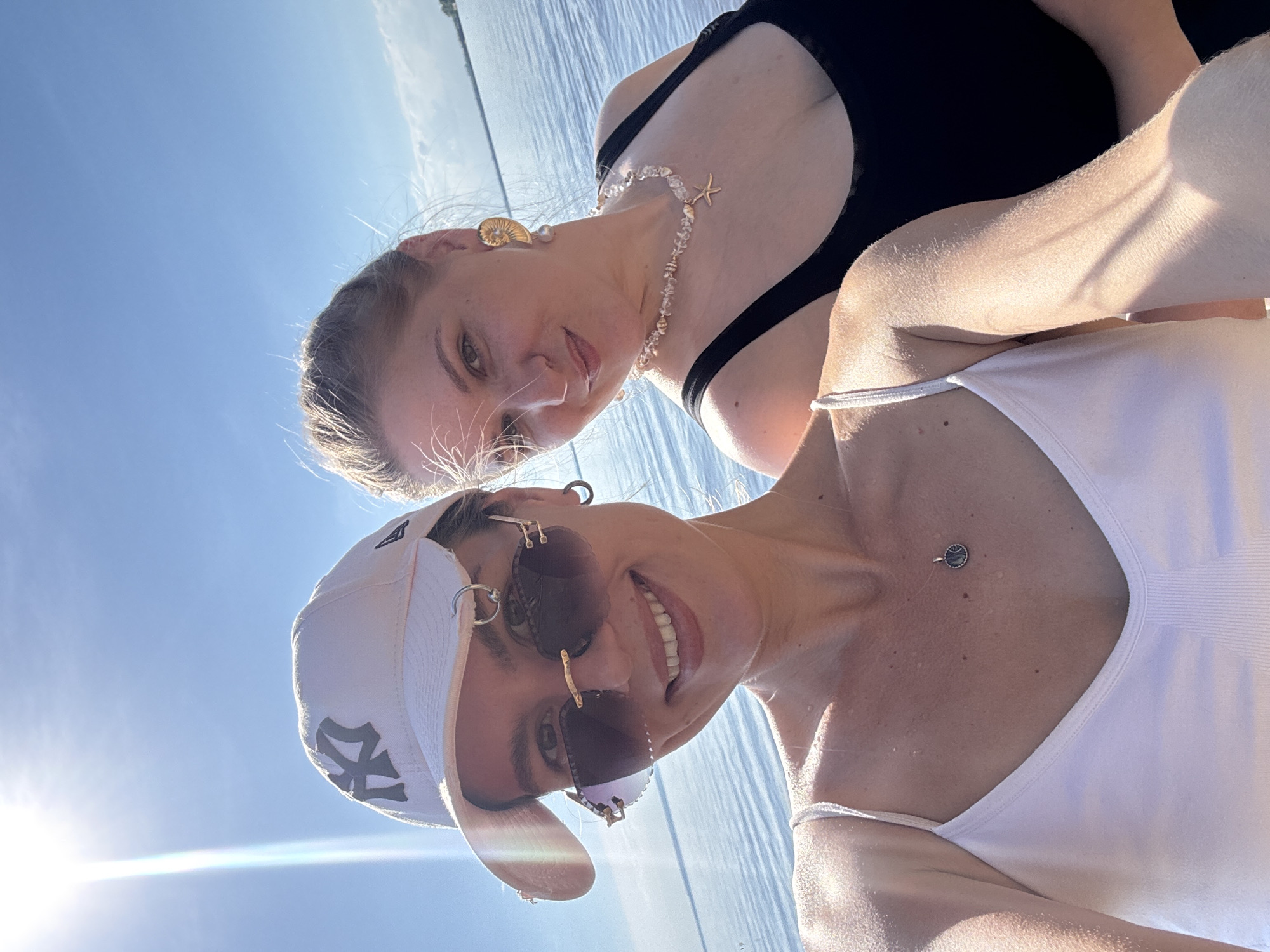 Two women are posing for a selfie outdoors with a bright sky and body of water in the background. The woman in the foreground is wearing a white tank top, a white baseball cap with a dark logo, and large, dark sunglasses. She has a necklace with a circular pendant visible. The woman behind her is wearing a black tank top and a necklace with seashell and starfish charms. She has large, golden earrings. The water appears calm and the sky is a clear blue with some faint clouds. The sun is shining brightly.