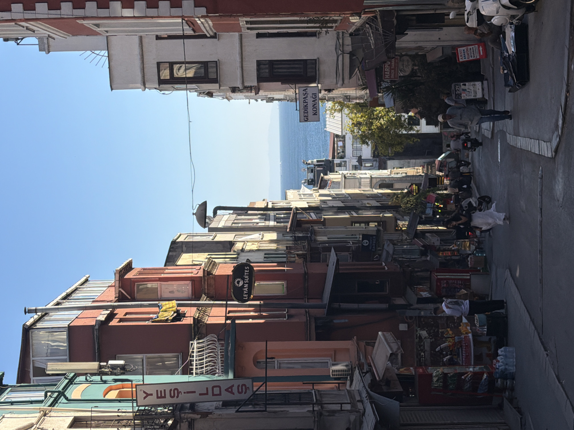 The image depicts a narrow, sloped street in the Balat neighborhood of Istanbul, Turkey, with buildings on either side. The buildings are colorful and have a mix of architectural styles, with some featuring balconies and bay windows. A clear blue sky is visible above.
In the foreground, a street vendor is selling goods from a cart. Several people are walking along the street, and a few motorcycles are parked. In the middle ground, signs for "LEYAN SUITES" and "GEDIKPAŞA KONAĞI" are visible. Further down the street, the sea is visible with a few boats on the water.
The overall impression is of a vibrant and historic urban environment, with a mix of residential and commercial buildings. The street appears to be a popular area, with a steady flow of people and activity. The sunlight suggests it is daytime, likely morning or afternoon.