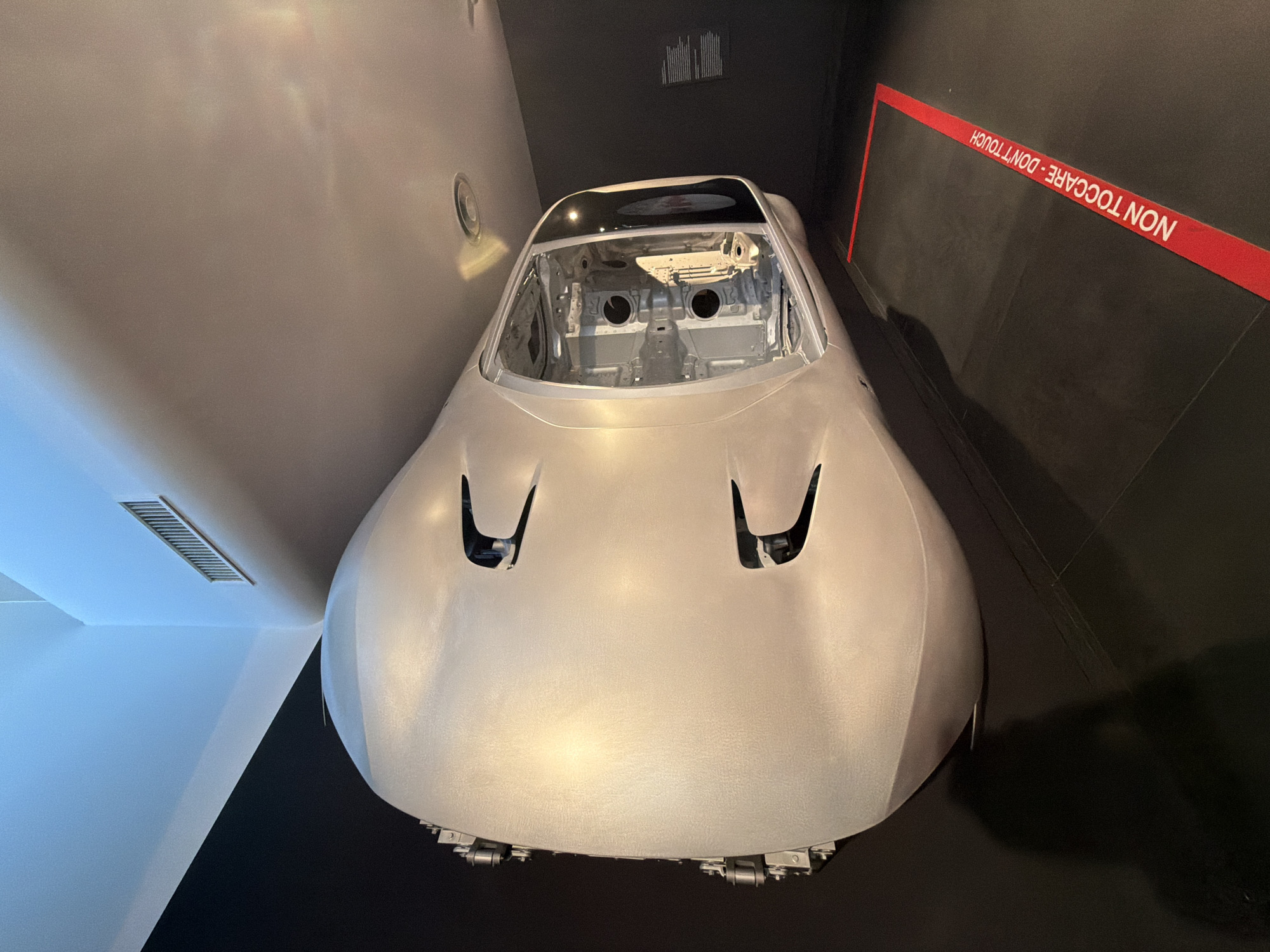 The image shows a silver, aerodynamic car chassis on display in a museum setting. The chassis is suspended or positioned in a way that suggests it's an exhibit, possibly in a factory or museum dedicated to automotive design or history. The interior of the chassis is exposed, revealing its internal structure. A red tape with the inscription "NON TOCCARE - DON'T TOUCH" is visible on the floor, indicating a restricted area around the exhibit. The background is dark with some lighting creating reflections on the car and ceiling. The scene is likely within the Ferrari Museum in Maranello, Italy, given the context of automotive exhibits and the Italian text on the floor.