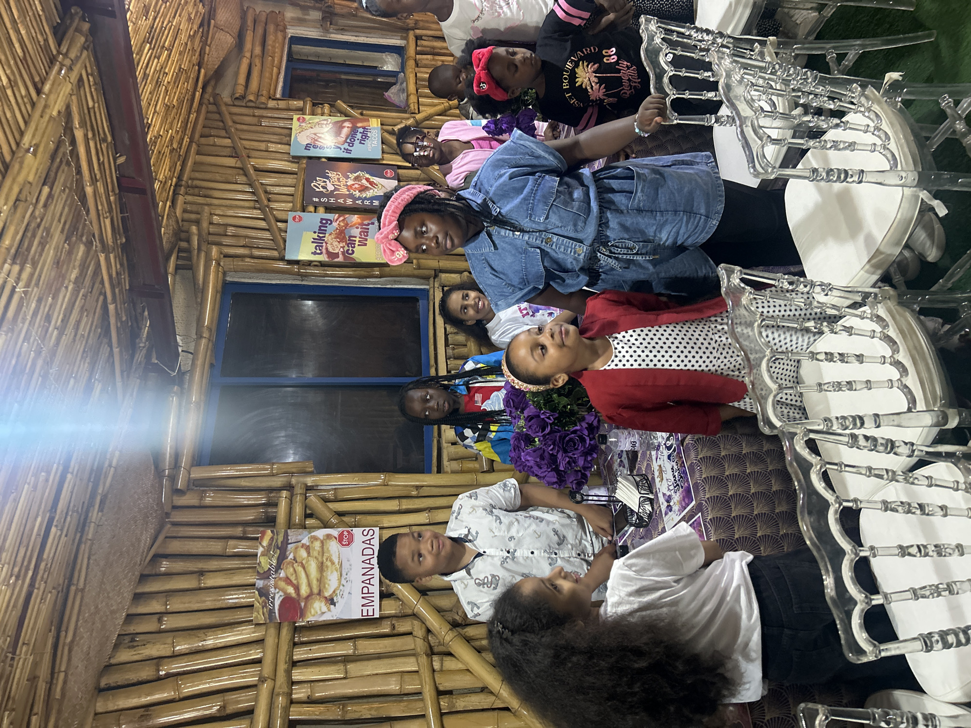 In Bar Jirgi Summa, Nigeria, a group of children are gathered indoors, likely at a restaurant or event space, with walls and ceiling constructed from bamboo. A poster advertising "EMPANADAS" is visible on the bamboo wall. The children are positioned around a table adorned with a purple patterned tablecloth. Several clear acrylic chairs with white cushions are arranged in front of the table. The children are of varying ages and appear to be interacting with each other. One young girl in the foreground is wearing a red jacket over a white polka dot dress and a pink headband. To her right, a girl in a denim jumpsuit and pink headband stands prominently. Other children are visible in the background, some looking towards the camera, others engaged in conversation. The lighting in the space is somewhat dim, with a bright light source emanating from the ceiling.