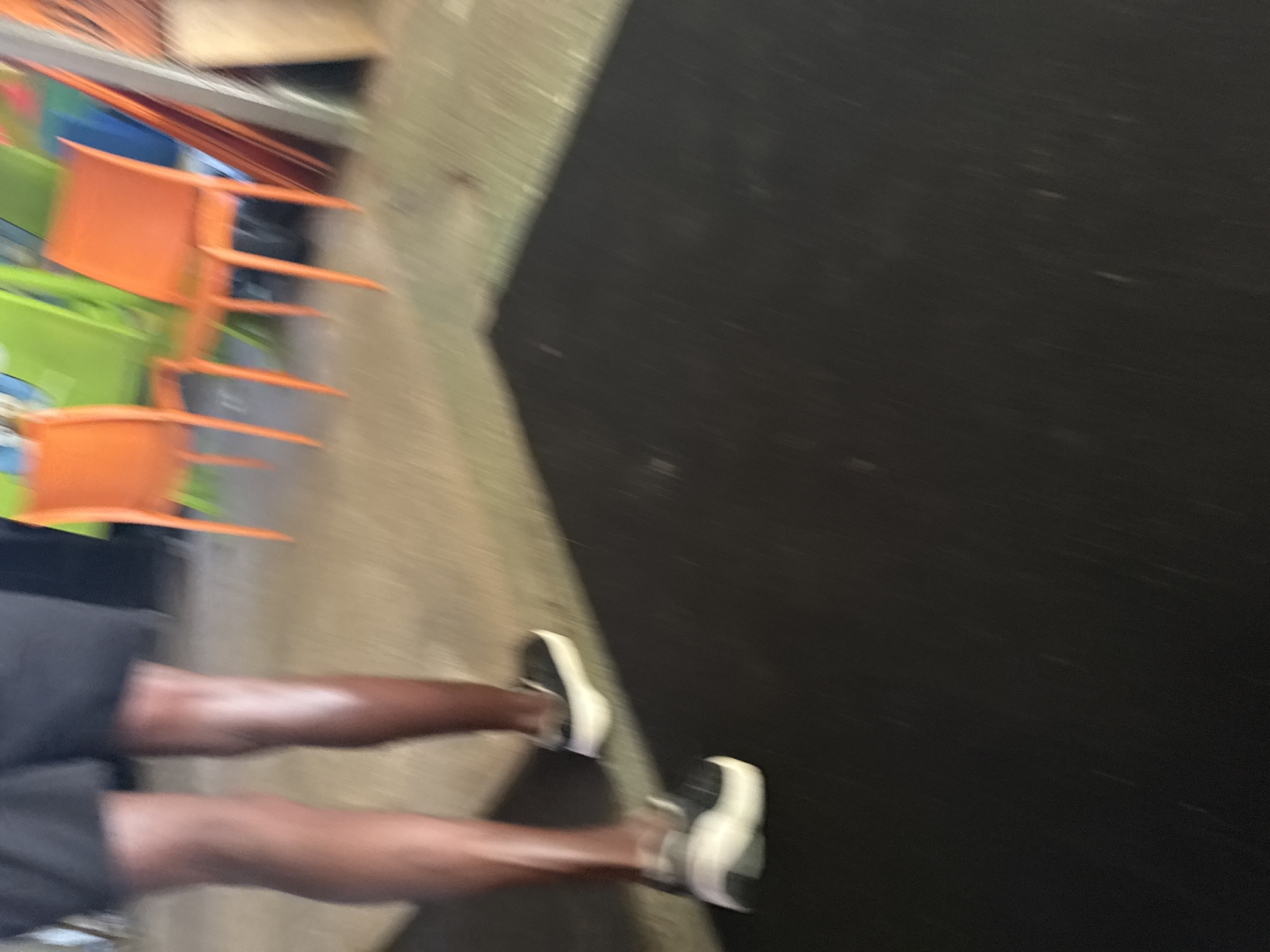 The image captures a close-up view of a person's legs and feet, wearing black shorts and white-soled sneakers with black uppers. The person is standing on a surface that transitions from a light-colored, possibly concrete or asphalt, area to a dark, mat-like surface. In the background, blurred out, are several bright orange chairs and some green objects, suggesting an outdoor or semi-outdoor setting possibly near a park or event space. The lighting is somewhat dim, and the motion blur indicates movement within the frame. There is no discernible indication of the city of Aksu, Turkey, in the image.