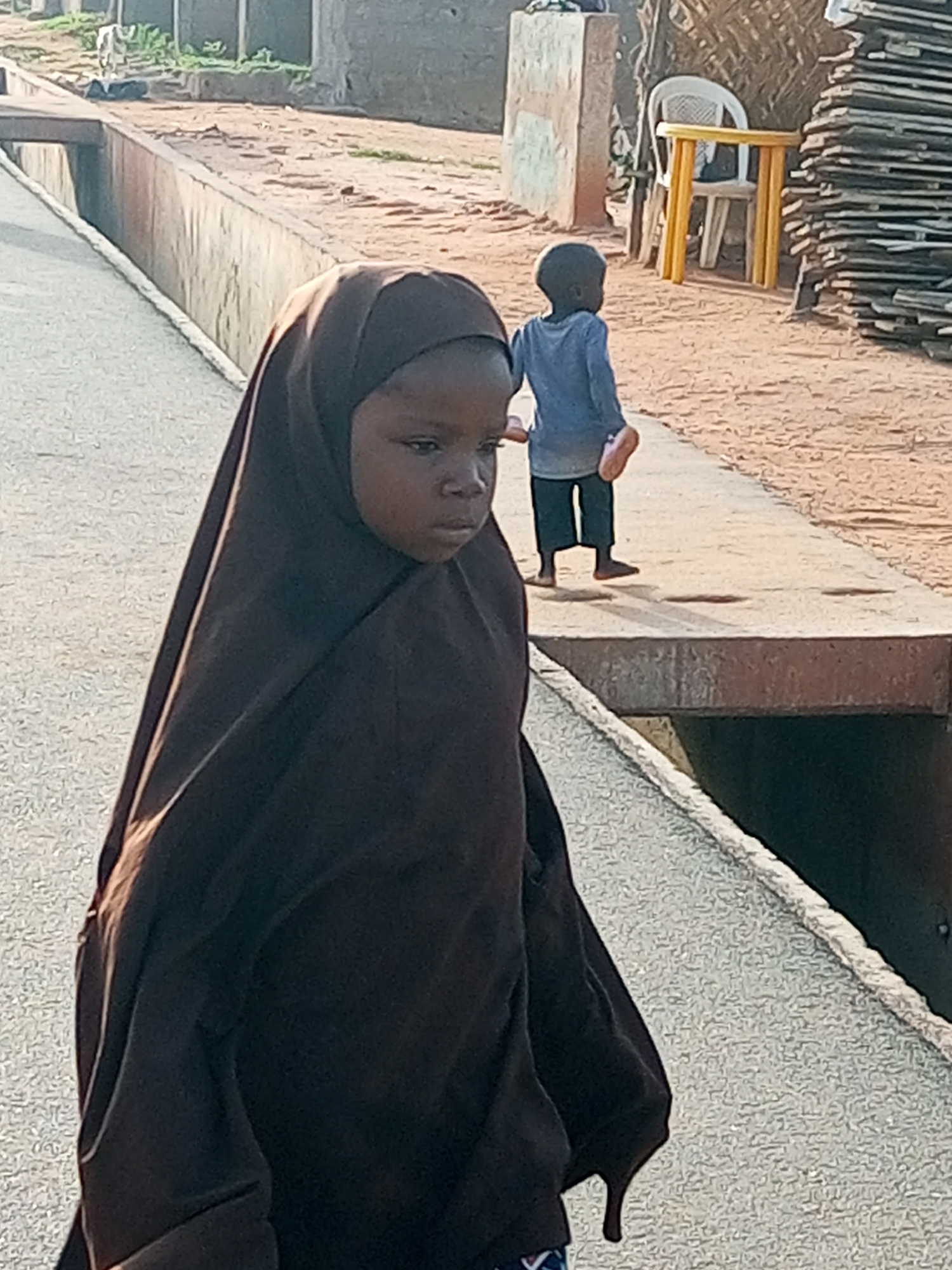 In the city of Gombe, Nigeria, on a dusty path next to a concrete drainage channel, two young children are observed. In the foreground, a young girl is wearing a dark brown hijab that covers her head and body. Her face is turned slightly to the side, and she appears to be looking down or away from the camera. Behind her, a younger child, possibly a boy, stands facing away from the viewer. He is wearing a blue long-sleeved shirt and dark pants. His feet are bare, and he is holding something light-colored in his right hand, possibly a shoe or sandal. In the background, a white plastic chair is positioned next to a yellow-legged table, with a stack of lumber or wood planks to the right. The environment appears to be an outdoor, possibly residential or communal area, characterized by dirt, concrete structures, and sparse vegetation. The lighting suggests it is daytime, with sunlight casting shadows.