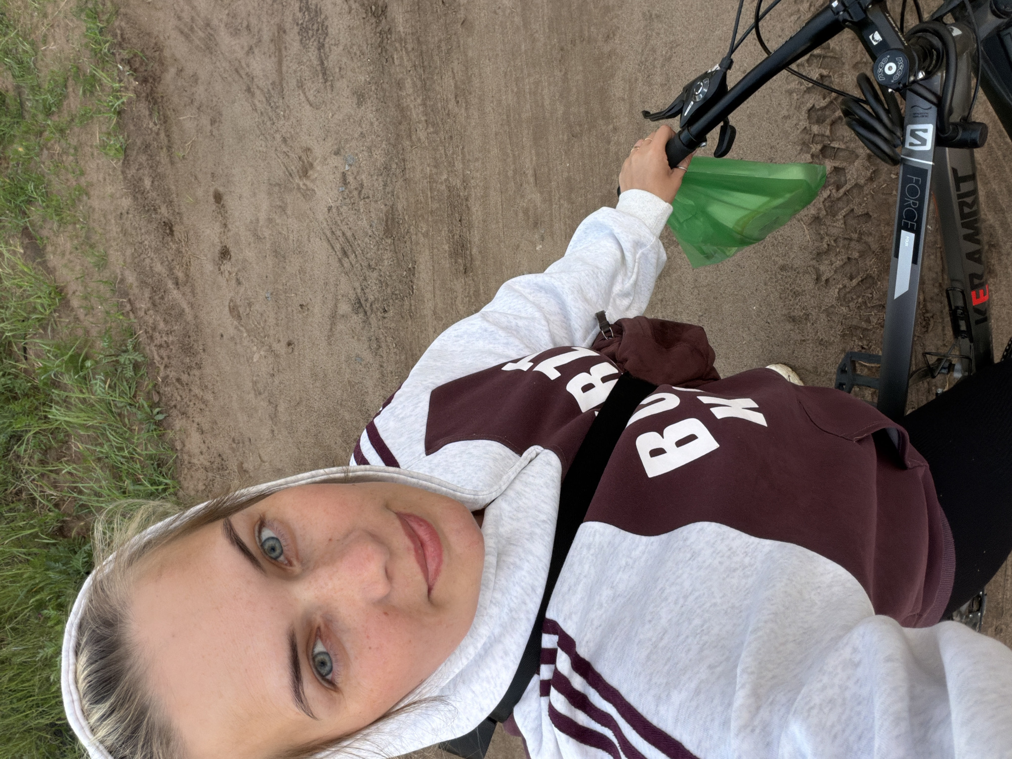A selfie taken from above shows a young woman with blue eyes sitting on a bicycle. She is wearing a hooded sweatshirt with a maroon and white design. Her face is visible, and she is looking towards the camera. The bicycle has black handlebars and a green plastic bag hanging from them. The ground is a mixture of dirt and grass. The location is not identifiable as Chaikovsky, Russia from the information available in the image.