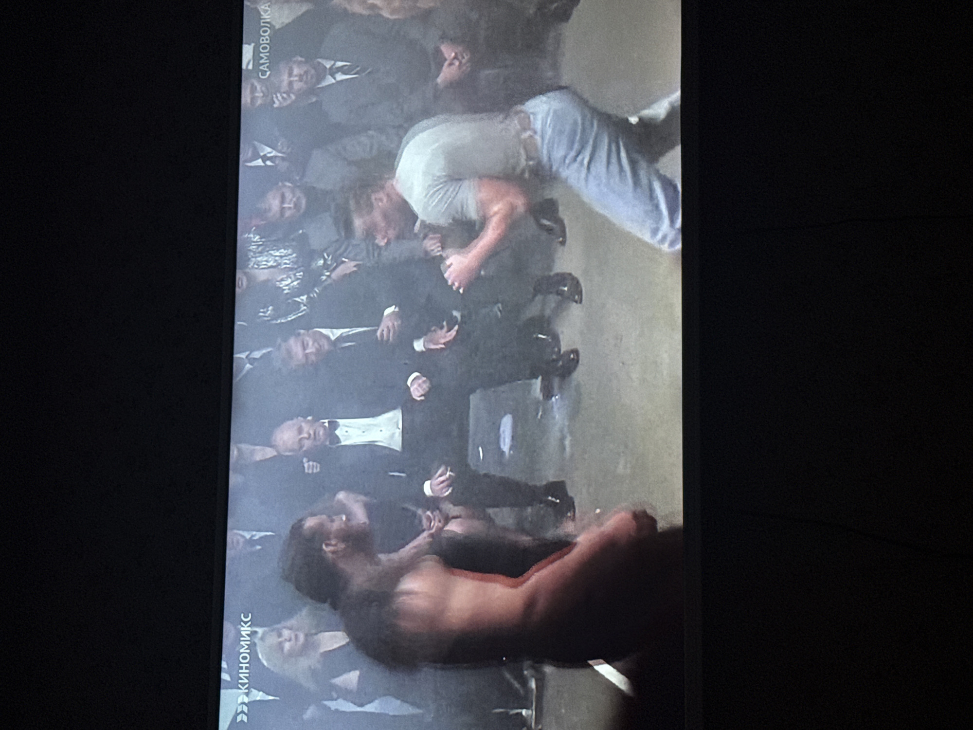A television displays a scene of violent conflict. A bare-chested man with long, dark hair grapples on the ground with another man wearing a light-colored t-shirt and blue jeans, seemingly in a bar fight amidst onlookers dressed in formal attire. The location, indicated by text in Cyrillic characters on the screen, is in Mytishchi, Russia.