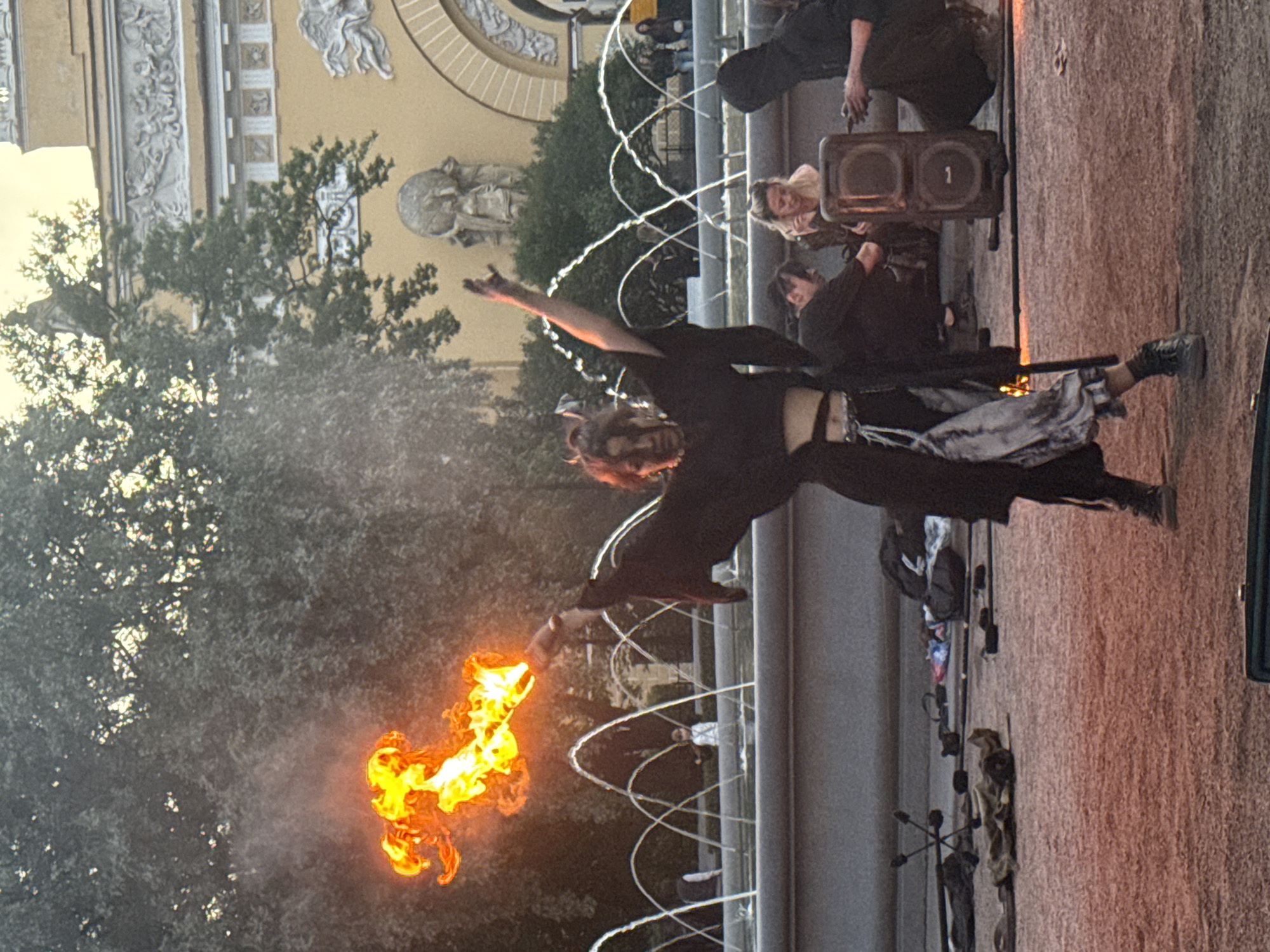 A person is performing with fire in front of a fountain in Palace Square, Saint Petersburg, Russia. The performer, dressed in black, is holding a flaming object in their right hand, with flames extending upwards. They are wearing a black robe and pants with a patterned skirt or wrap. Several other people are visible in the background, some sitting and watching, and others standing near a large speaker. The architecture of the General Staff Building is visible in the background, with its yellow facade and classical ornamentation. The scene is set during the day, with soft lighting and shadows.
