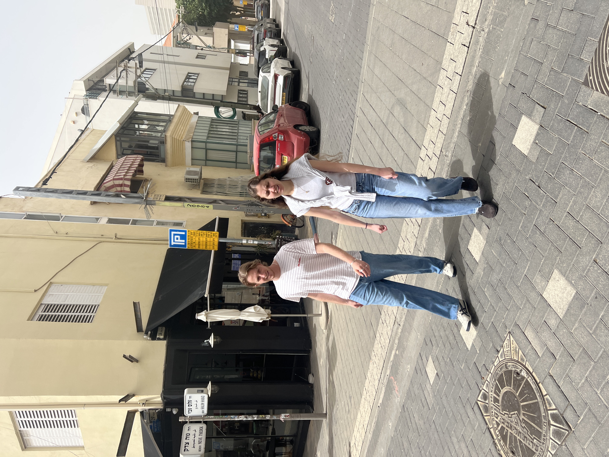 The image shows two people standing on a brick-paved street in Tel Aviv, Israel. Both are wearing similar clothing: blue jeans and white shirts. The architecture surrounding them includes buildings with light-colored facades and awnings. Street signs are visible with Hebrew and possibly Arabic inscriptions. A red car and other vehicles are parked along the street. A manhole cover with inscriptions is visible on the ground. The street signs indicate they are at the intersection of Rehov Neve Tzedek and Rehov Shalom Shabazi. A sign for parking restrictions is seen as well.