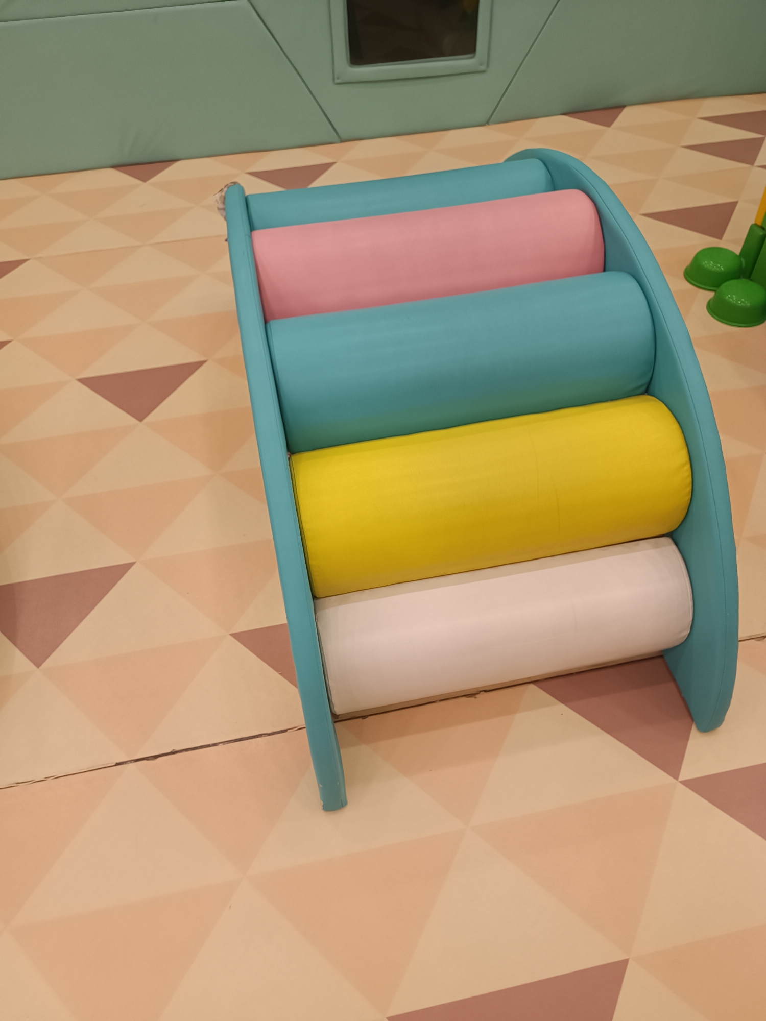 The image shows a children's play structure with four cylindrical cushions: pink, turquoise, yellow, and white. The structure is light blue and is placed on a floor with a geometric pattern of light and dark pink triangles. In the background, there is a light green wall and a dark rectangular mirror. To the right of the play structure, there are two green, mushroom-shaped toy pieces. The scene appears to be indoors, likely in a playroom or kindergarten. There are no people visible in the image. This scene is not located in Baku, Azerbaijan, as there are no visual cues to suggest that location.