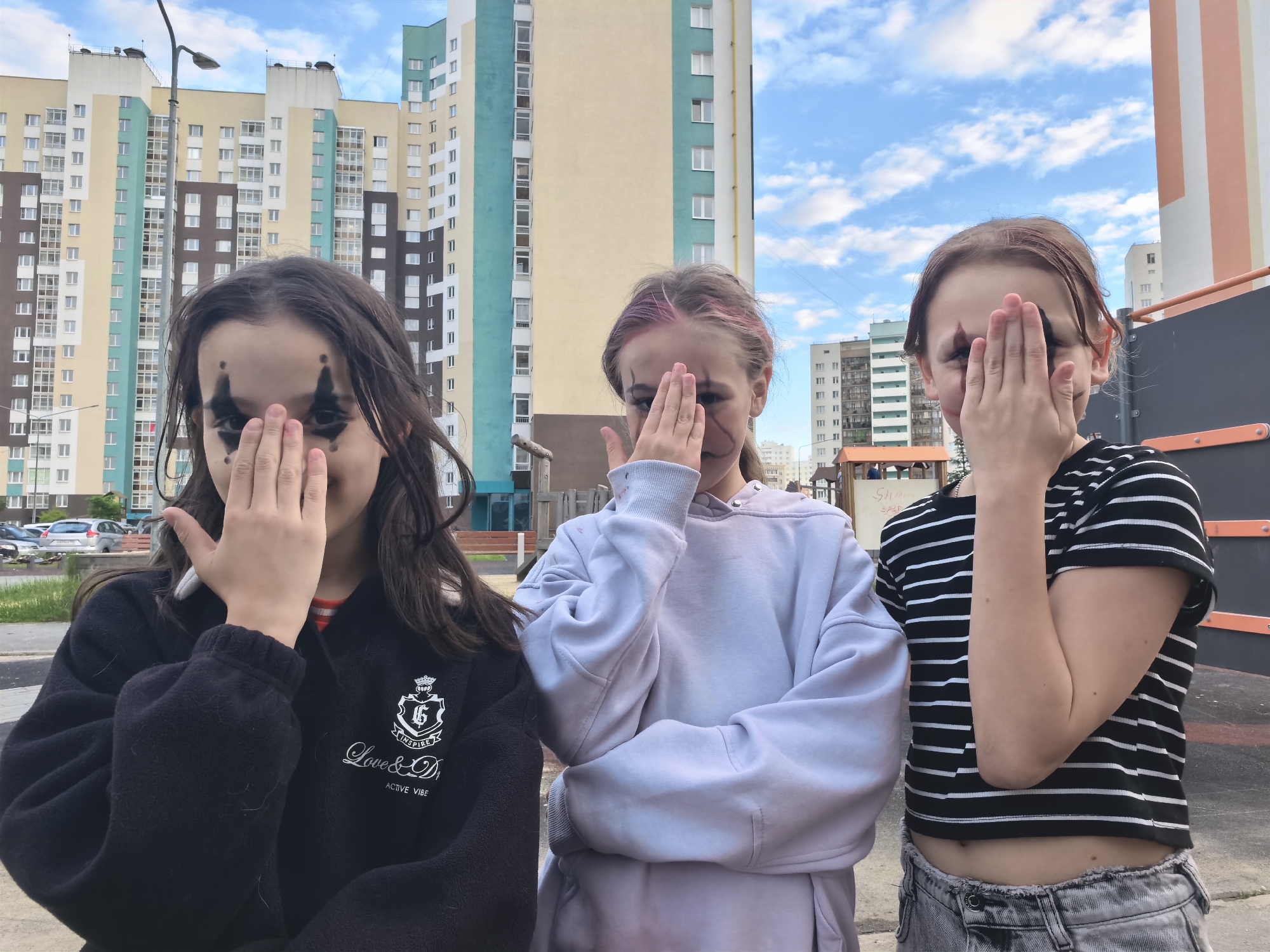Three young females stand in front of a building complex with a cloudy blue sky above. The female on the left has dark hair and black makeup around her eyes, obscuring her face with her hand. She wears a black fleece jacket with white text and graphics. The female in the middle has brown hair with pink streaks, and she covers her face with her hand. She is wearing a light purple hooded sweatshirt. The female on the right has brown hair and wears a black and white striped t-shirt and distressed gray jeans, also covering her face with her hand. The environment appears to be an urban outdoor space with buildings, a lamppost, a grassy area, and a playground structure visible in the background.