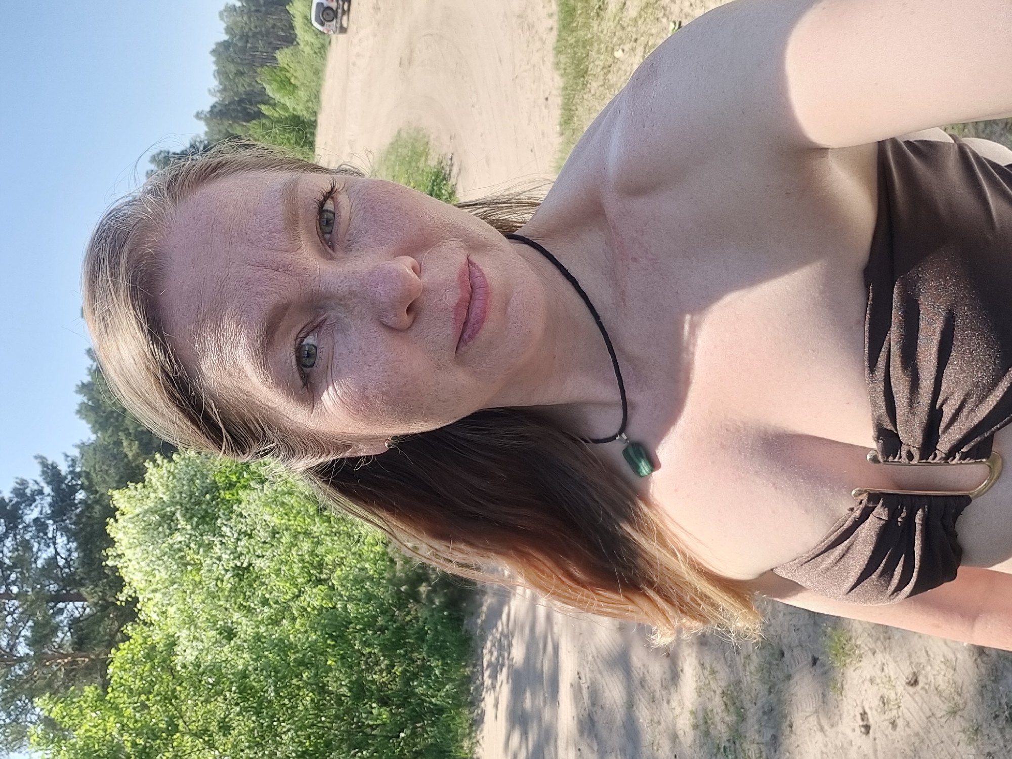 A woman is outdoors, positioned on a sandy path or road with trees and green foliage visible in the background. She is fair-skinned, with freckles on her face, and has light brown hair. She is wearing a brown, strapless top with a metal buckle detail, and a black necklace with a green pendant. A white vehicle can be seen in the distance. The location is Chaikovsky, Russia.