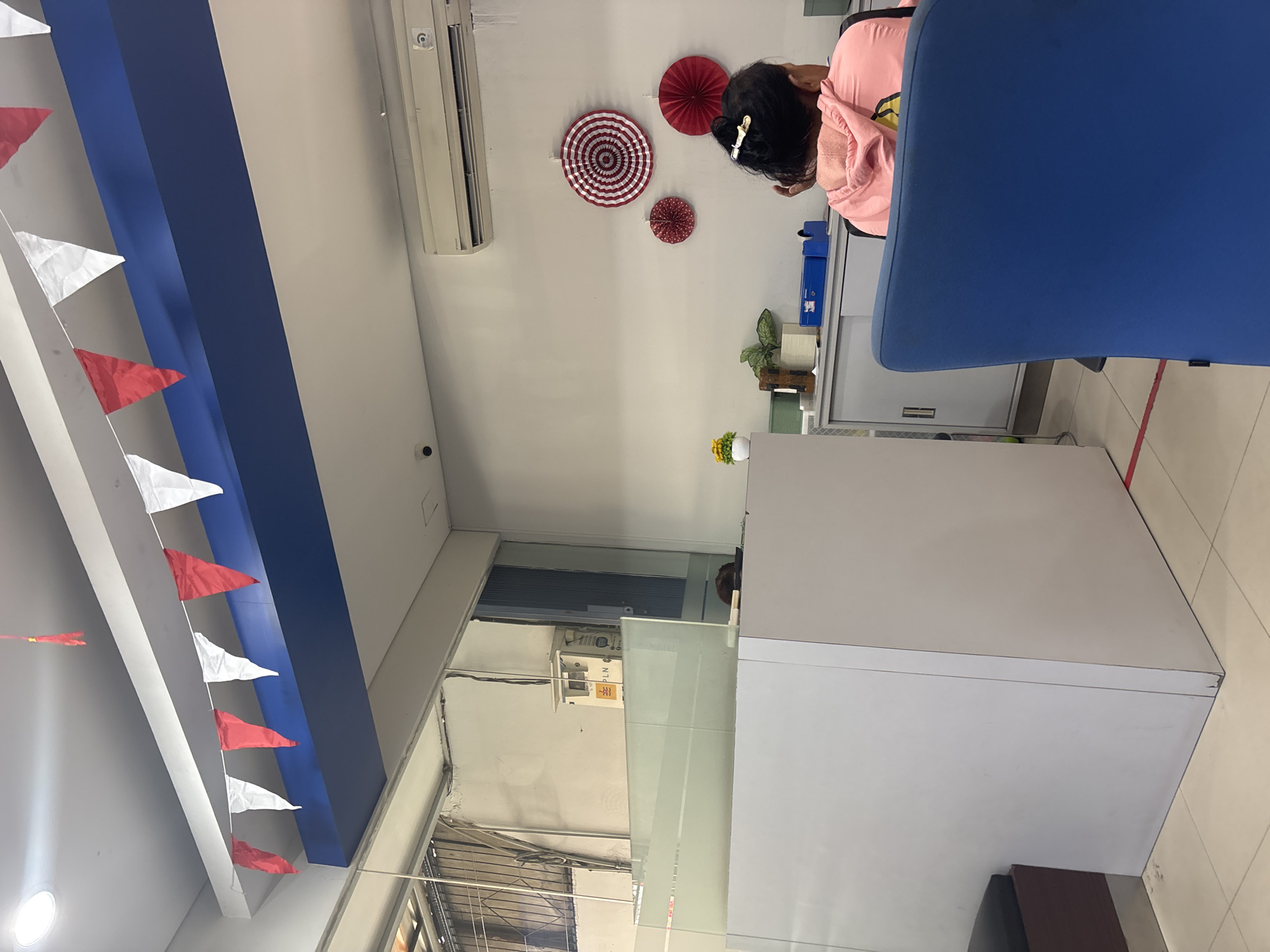 The image depicts an indoor scene, likely an office or service area, within North Jakarta, Indonesia, as evidenced by the "PLN" signage on a meter box, which refers to Perusahaan Listrik Negara, the state electricity company of Indonesia. In the foreground, a large, light-colored desk or counter occupies the left side of the frame, with a dark-colored seating area to its left. A person, viewed from the back, is seated in a bright blue chair on the right, wearing a pink hooded jacket. They have dark hair styled with a clip. The background reveals a wall adorned with red and white circular decorations and an air conditioning unit. A security camera is visible on the ceiling. A string of red and white triangular flags is hung across the upper portion of the room. The environment appears to be a functional space with administrative elements like cabinets and office supplies.