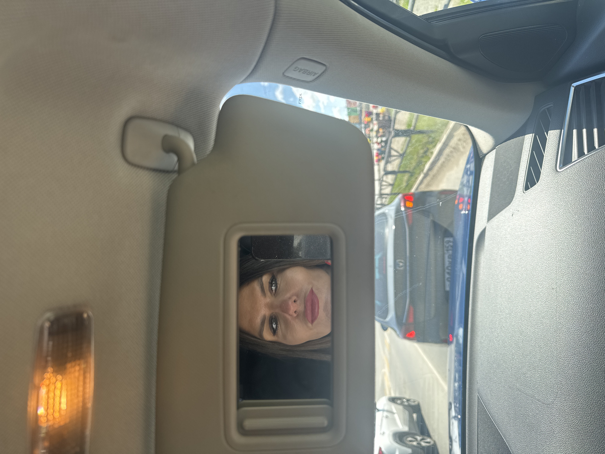 The image shows a woman's reflection in a car's visor mirror. She is looking slightly to her left. The woman has brown hair, makeup on her face, and is wearing a red top. The car is in motion, with other vehicles visible in the background, suggesting it is on a road. The interior of the car is visible, including part of the dashboard and the visor itself, which has an overhead light. The scene appears to be a daytime shot, with sunlight streaming in. The location of Chaikovsky, Russia, cannot be determined from the visual information provided in the image.