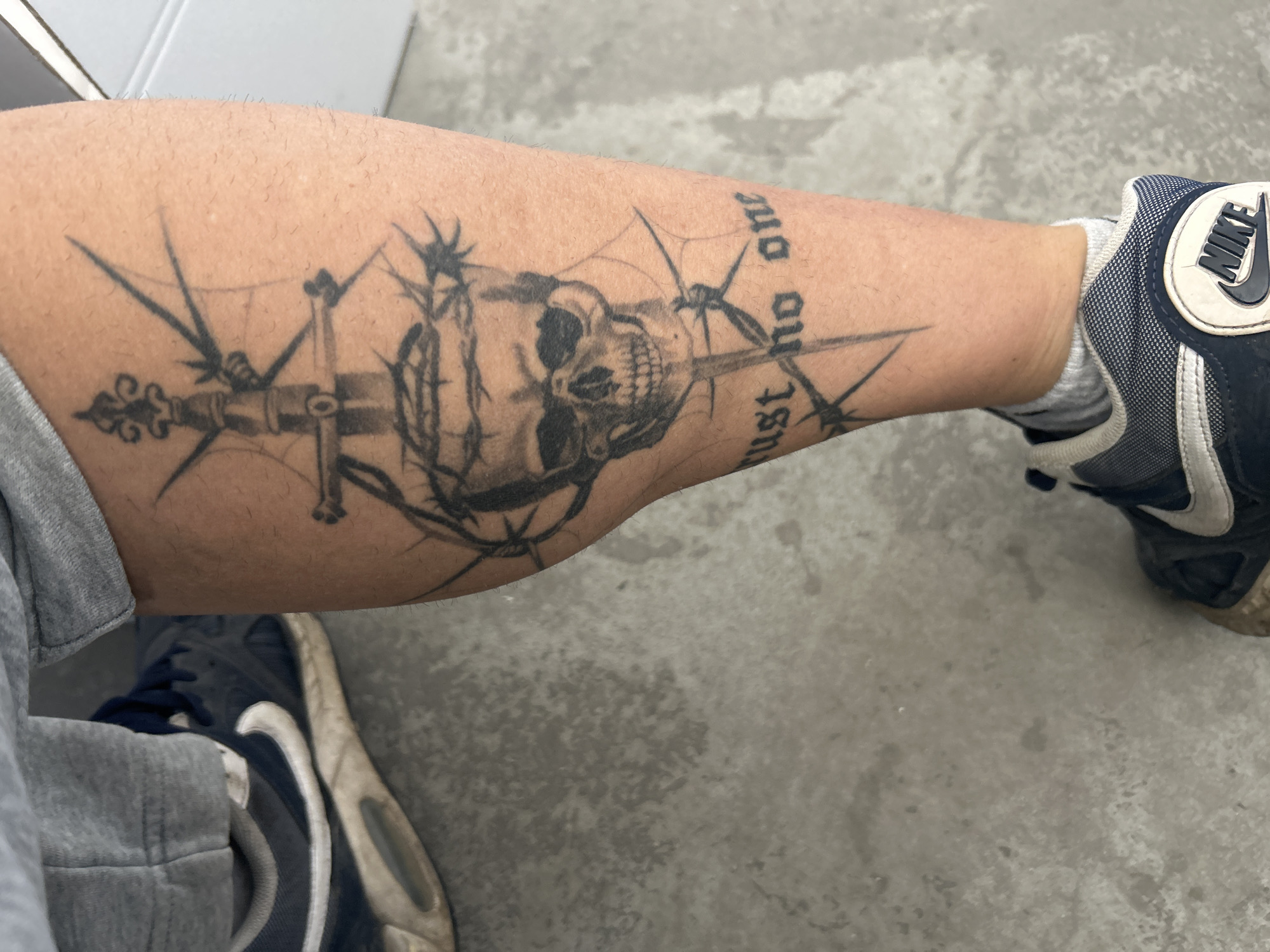 The image shows a person's lower leg with a tattoo featuring a skull, a sword, and barbed wire. The tattoo is black and gray, and includes the text "trust no one." The person is wearing a gray sweatpants leg and a blue and white Nike sneaker. The background is a gray concrete floor.