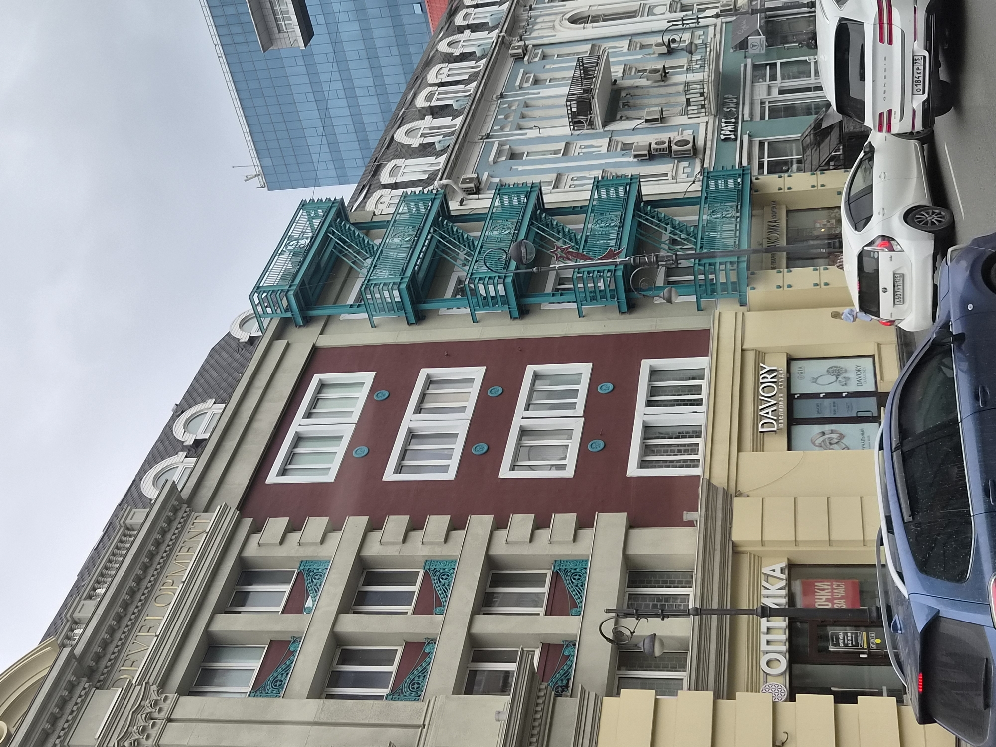 This image captures a street scene in Vladivostok, Russia, showcasing a blend of architectural styles. In the foreground, a dark blue car is partially visible, facing away from the camera. Further down the street, a white hatchback and a white SUV are parked.
The buildings lining the street present a varied facade. To the left, a light-colored building with decorative elements and repeating window patterns stands adjacent to a maroon-colored building with similar window designs. A prominent teal-colored external staircase ascends the side of the maroon building.
Further along, a light blue building with ornate architectural details and balconies is visible. Behind these buildings, a modern, blue glass skyscraper rises, contrasting sharply with the older structures. Various shop signs are present on the ground floors of the buildings, including "OPTIKA" and "DAVORY". The sky is overcast, suggesting a cloudy day.