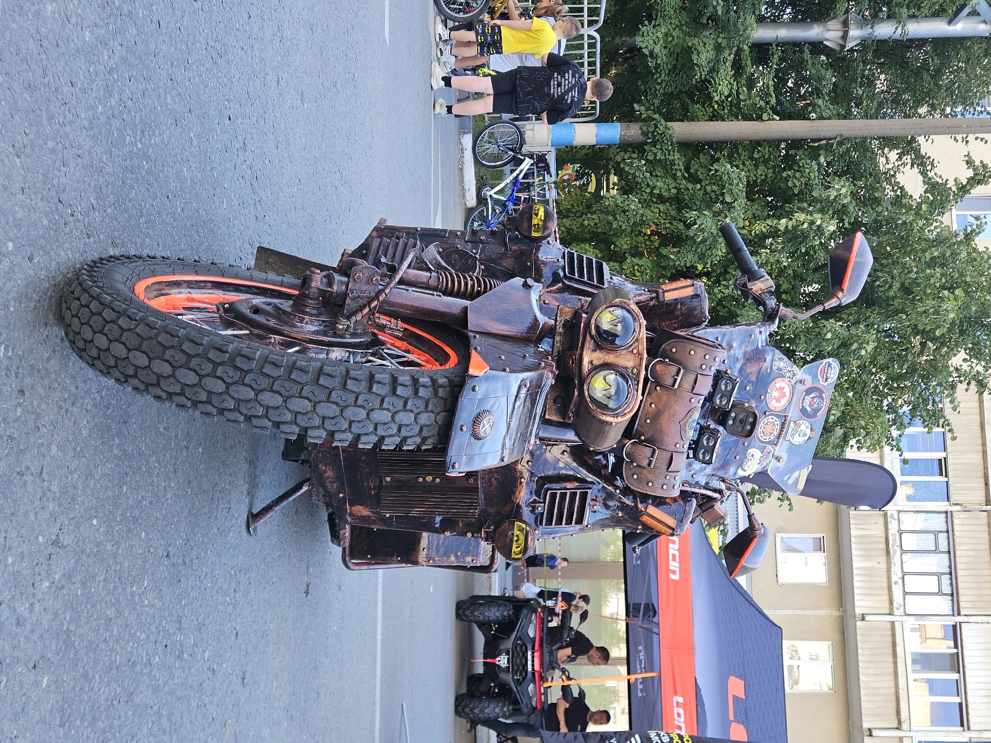 A custom-built, heavily modified motorcycle with a weathered, metallic brown finish and intricate detailing stands prominently on an asphalt street in Ulyanovsk, Russia. The motorcycle features dual headlights, prominent leather saddlebags, and a windshield adorned with numerous stickers. Its front tire, with an orange rim, is large and rugged. To the left of the motorcycle, a group of individuals, including a boy in a yellow t-shirt and another in a black t-shirt, are visible. In the background, a building with multiple windows and trees can be seen. To the right, an orange tent with red lettering and an all-terrain vehicle (ATV) are present, with two men standing near the ATV. The scene appears to be an outdoor event or exhibition.