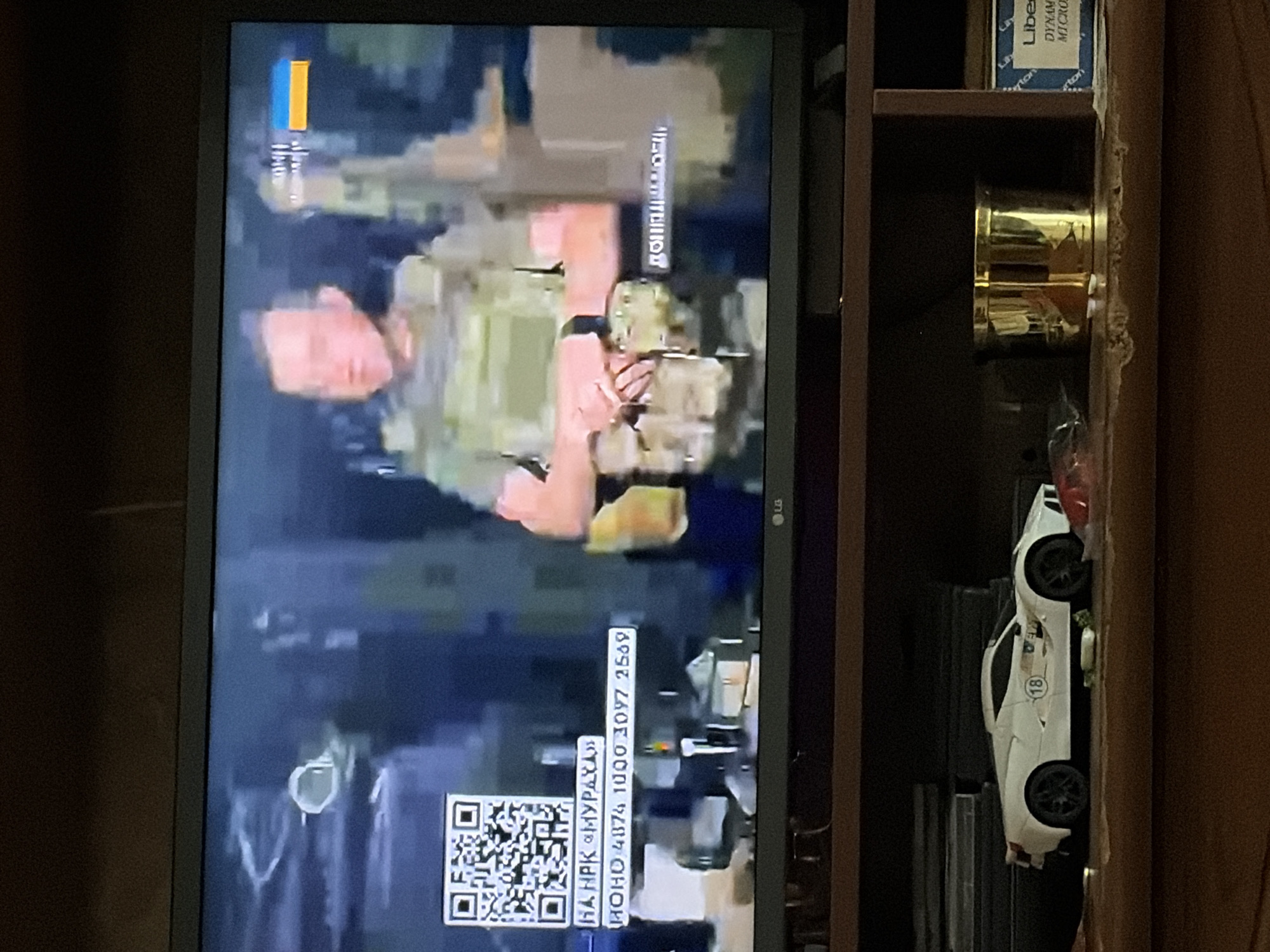 The image shows a television screen displaying a news report. A man wearing tactical gear is visible on screen, with a Ukrainian flag icon in the upper right corner. Text overlays on the screen include a QR code and what appears to be a donation account number. Below the television, a shelf contains a white toy sports car with the number 18 on it, a golden cylindrical object, and a box with text that is partially obscured but appears to be for "Liberty" and "Dynami Micro." The background of the image is a dark, indoor environment.