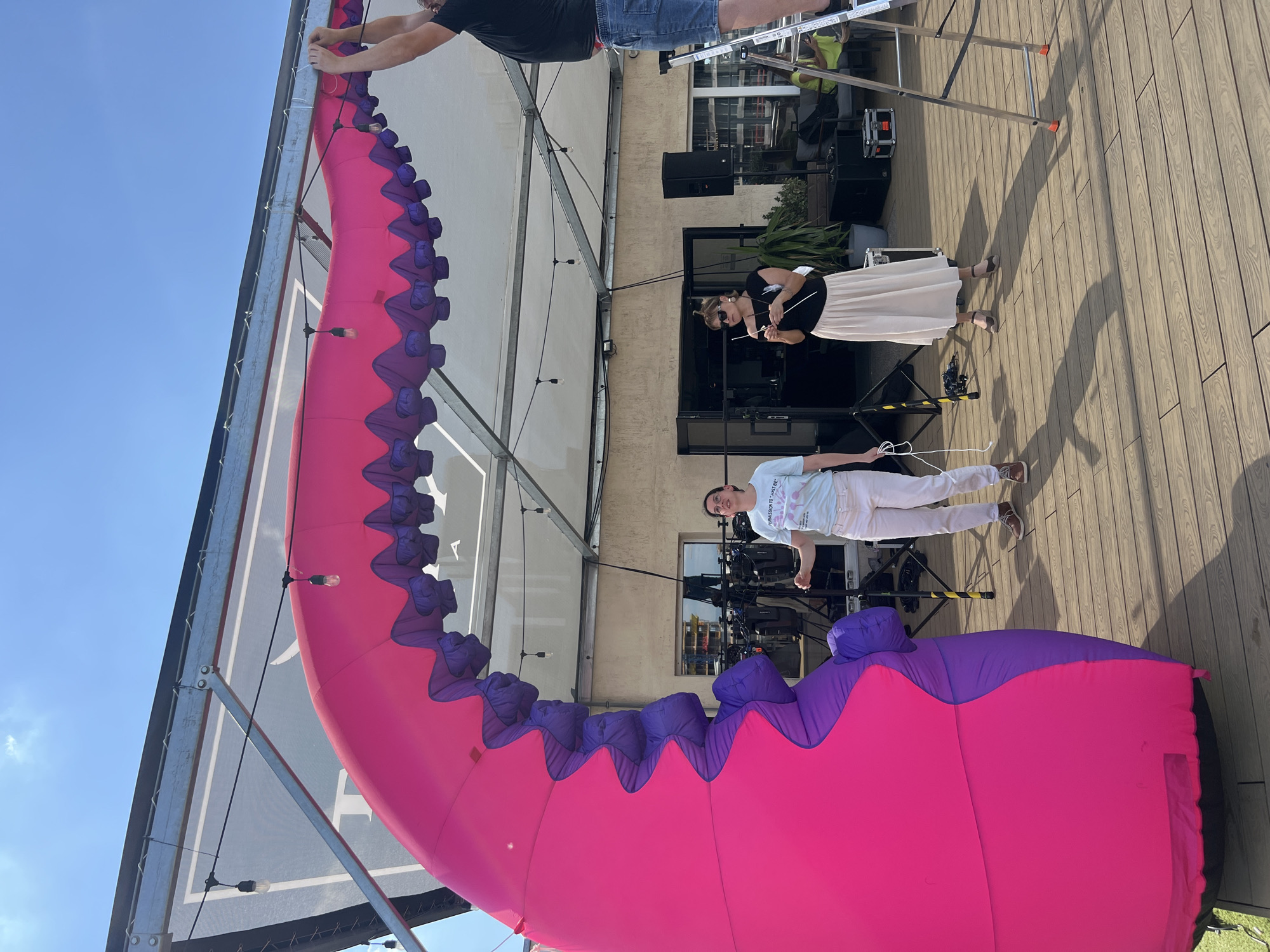 An inflatable tentacle-shaped decoration, predominantly bright pink with purple ruffled edges, is being installed outdoors. The structure is suspended from a metal framework covered by a translucent white canopy. Three individuals are involved in the installation. On the left, a woman wearing a light blue t-shirt and white pants stands near the base of the tentacle, holding a white rope. In the center, a woman in a black top and a long, flowing beige skirt holds a white stick or pole. On the right, a person wearing a black t-shirt and denim shorts stands on a silver ladder, adjusting the top of the tentacle. The scene takes place on a wooden deck, with various technical equipment, including speakers and stands, visible in the background. The sky is clear blue. This event is taking place at the rooftop of the National Palace of Culture (NDK) in Sofia, Bulgaria.