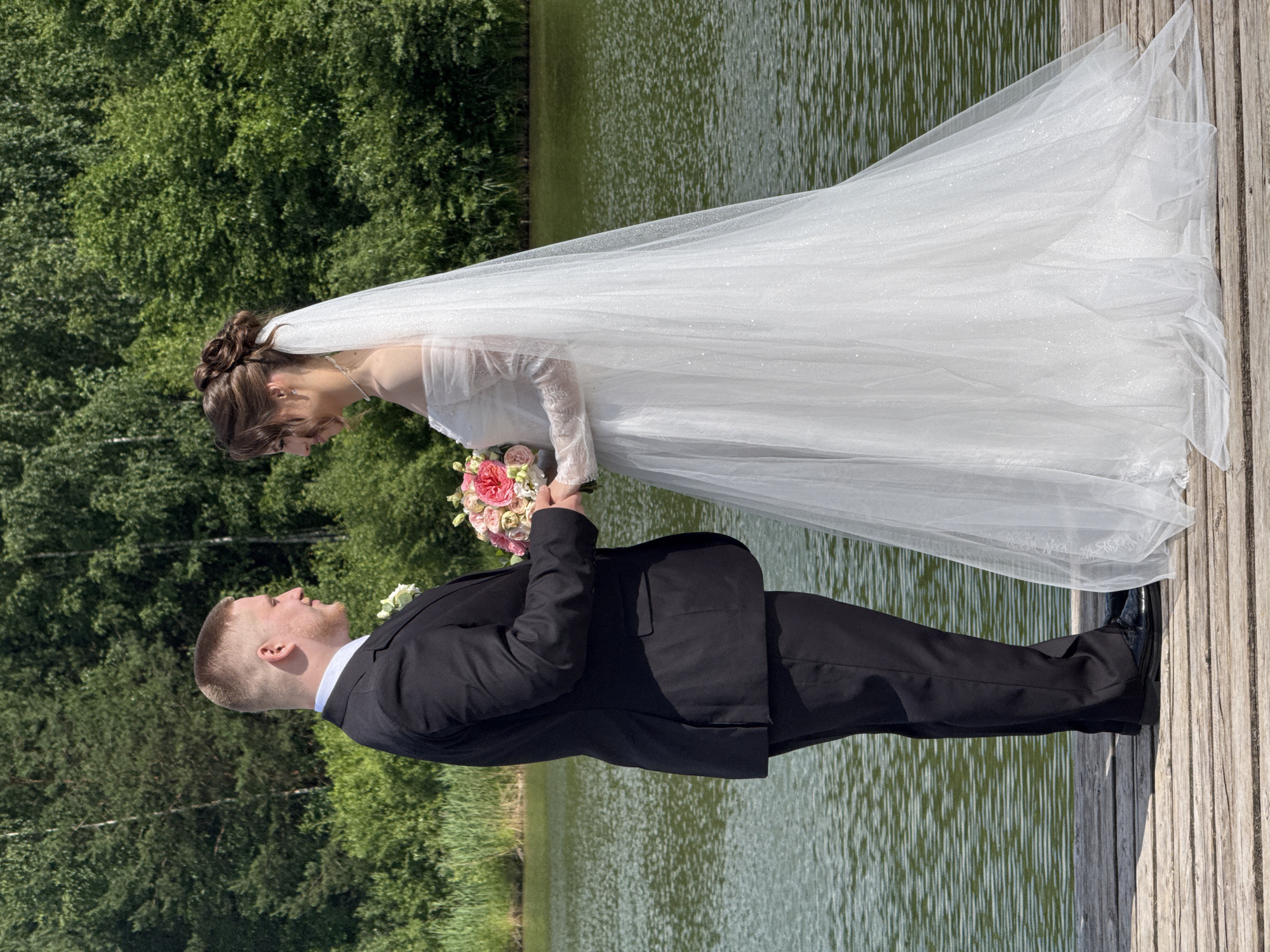 A full-body shot captures a newlywed couple standing on a wooden dock by a body of water. The groom, on the left, is dressed in a black suit, white shirt, and black tie. He is holding a bouquet of pink and white flowers in his left hand. The bride, on the right, wears a white off-the-shoulder wedding gown with long lace sleeves and a sparkling veil. She is looking at the groom and smiling. The water behind them is calm and green, reflecting the surrounding lush green trees. The wooden dock has visible planks. The scene appears to be set in a natural, outdoor environment, possibly a park or a lakeside area in Votkinsk, Russia, given the backdrop of trees and water.