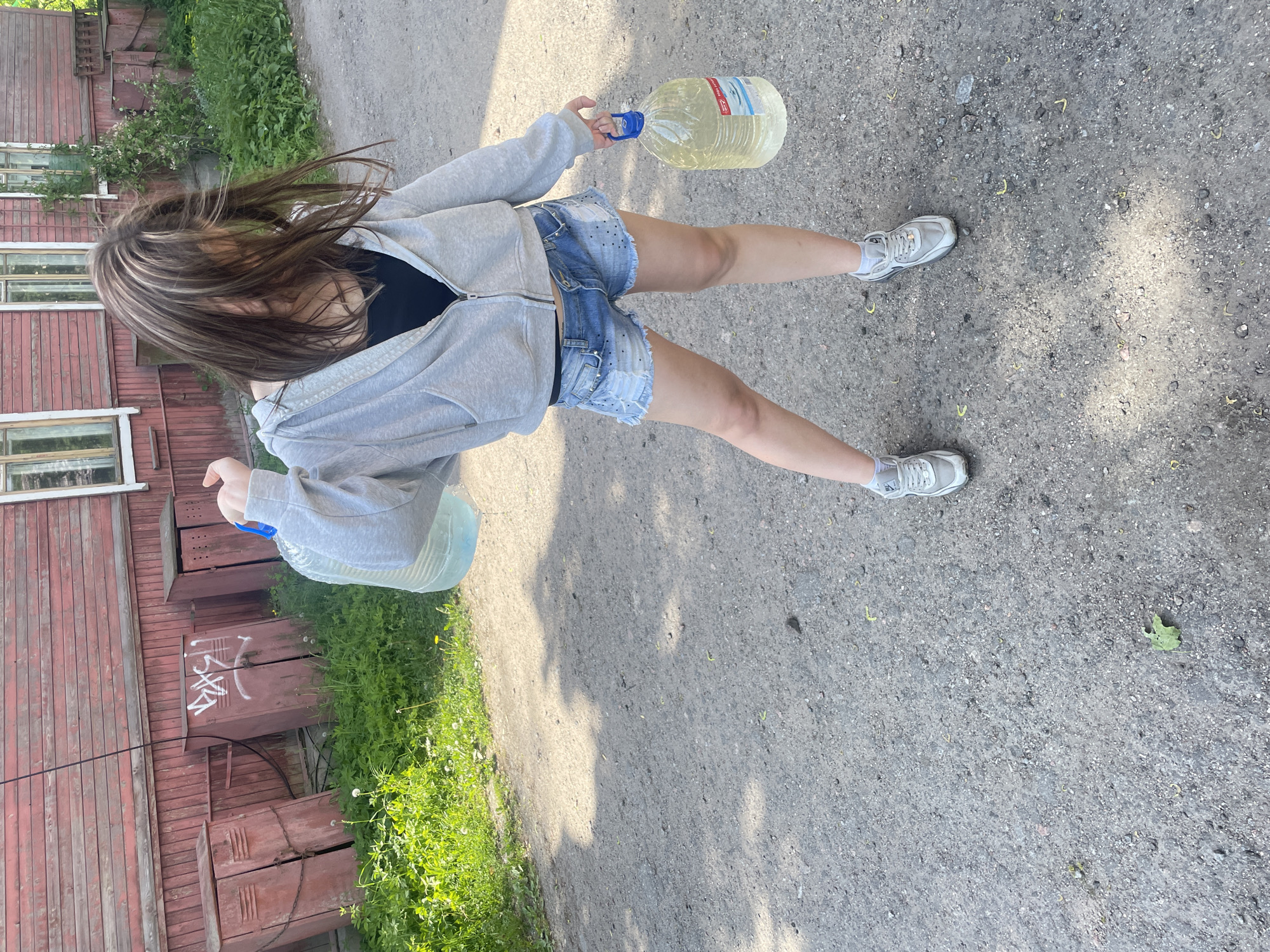 The image depicts a young adult female carrying two large plastic bottles of clear liquid on a gravel path in Priozersk, Russia. The female, with long brown hair featuring lighter streaks that obscure her face, is dressed in a grey hooded zip-up sweatshirt over a black top, distressed light blue denim shorts, and grey and white athletic shoes (sneakers) with white socks. She carries one bottle, equipped with a blue handle, slung over her left shoulder and resting against her back, while holding a second identical bottle by its blue handle in her right hand.
The ground is a coarse grey-brown gravel and dirt path, interspersed with small stones and minor organic debris, exhibiting distinct shadows indicative of direct sunlight. To the left of the path, a strip of vibrant green vegetation, including grass, weeds, and dandelions, runs along the base of a reddish-brown wooden building. This building features horizontal clapboard siding and multiple white-framed windows. Positioned against the building, partially obscured by the ground-level vegetation, are three weathered, reddish-brown metal utility boxes or cabinets, each with visible hinges and latches; the central box bears white graffiti spelling "ВАСЯ". A dark cable or wire is visible diagonally spanning the upper left portion of the background.
