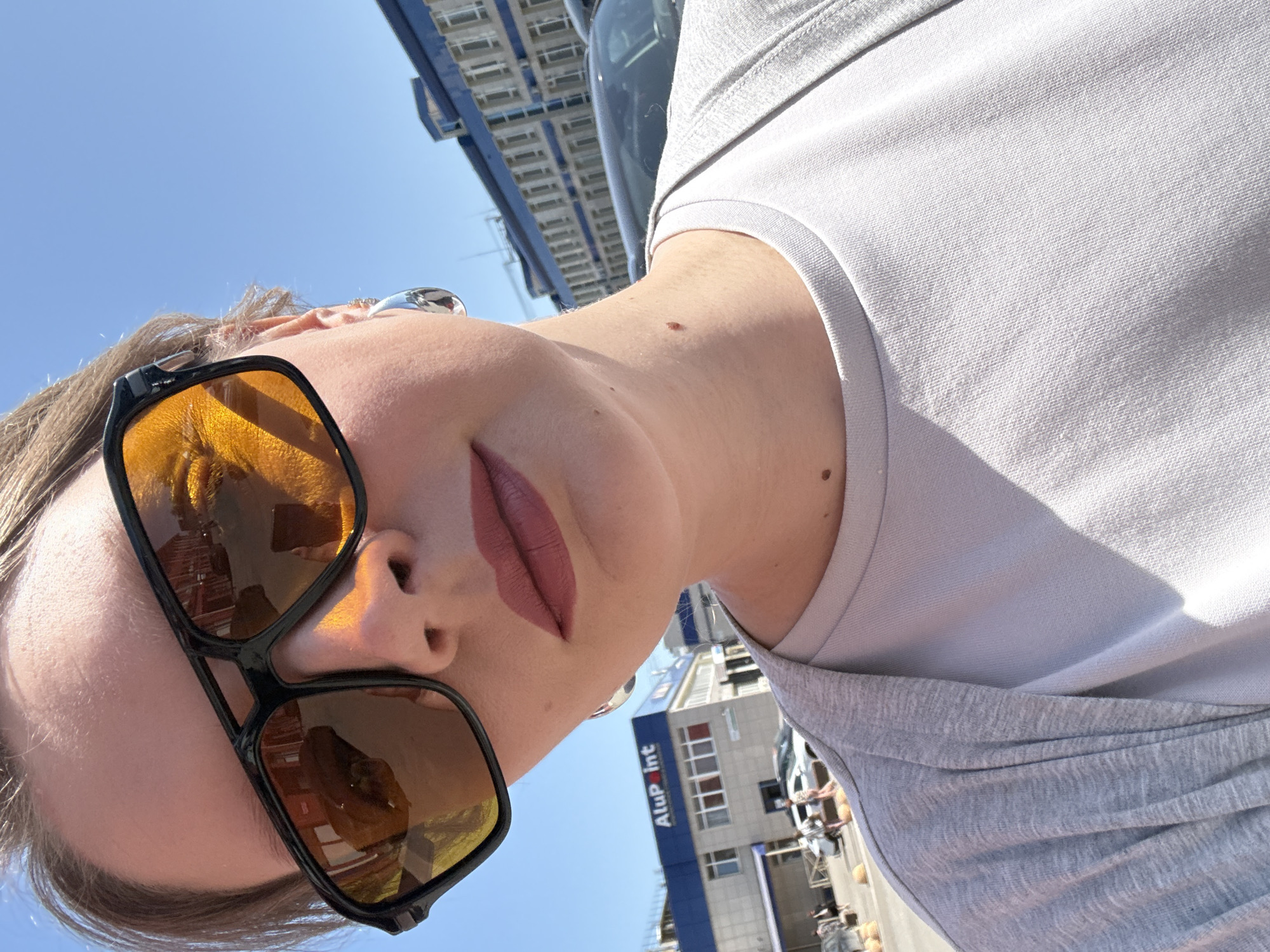 A low-angle close-up shows a person wearing sunglasses with orange lenses and a light-colored shirt. The background reveals a building with "AluPoint" signage and construction scaffolding, placing the location in Izhevsk, Russia. The sky is clear and blue. The person's face is partially visible, showing lips with mauve lipstick.