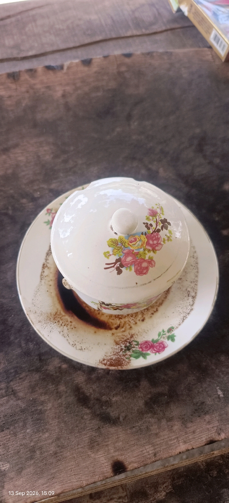 A covered ceramic bowl with floral decorations sits on a saucer with a matching floral pattern, placed on a weathered wooden surface. The saucer is stained with dark residue, likely from coffee grounds, which have spilled onto the wood. The background shows more of the textured wooden surface and a sliver of a cigarette pack. The timestamp "13 Sep 2026, 15:09" is visible in the lower left corner. The scene appears to be outdoors, given the natural light and rustic setting, consistent with a location in Mojopetung, Indonesia. No individuals are present in the image.