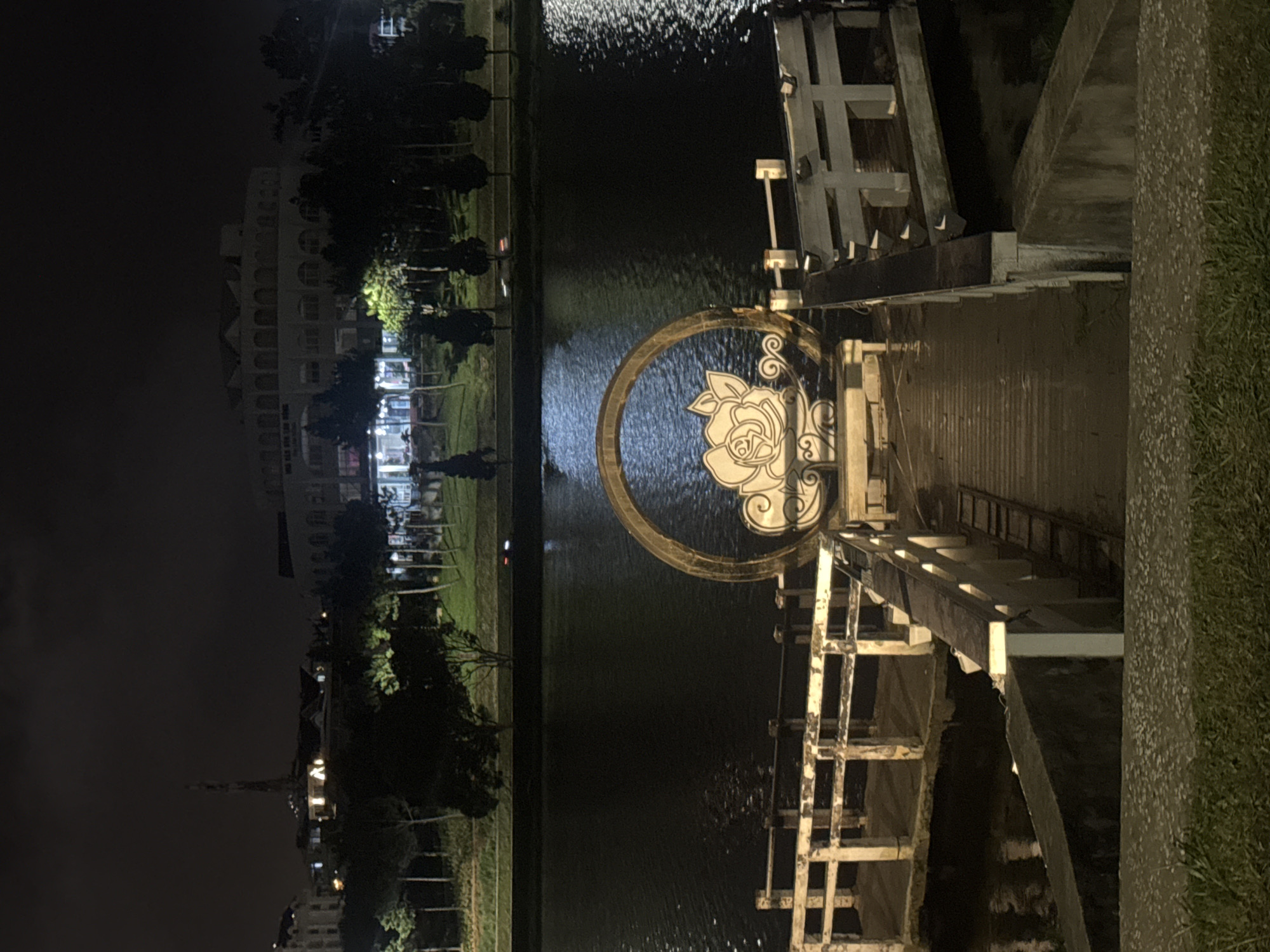 A nighttime photograph captures a waterfront promenade with a circular decorative arch featuring a white rose design. The arch is positioned at the end of a wooden pier that extends into a dark body of water. The pier is lined with white railings and has a wooden walkway. In the background, across the water, is a city skyline illuminated at night, with buildings and trees silhouetted against the dark sky. A tall, slender tower is visible to the left of the skyline. The overall scene depicts a serene, illuminated waterfront area in Da Lat, Vietnam.